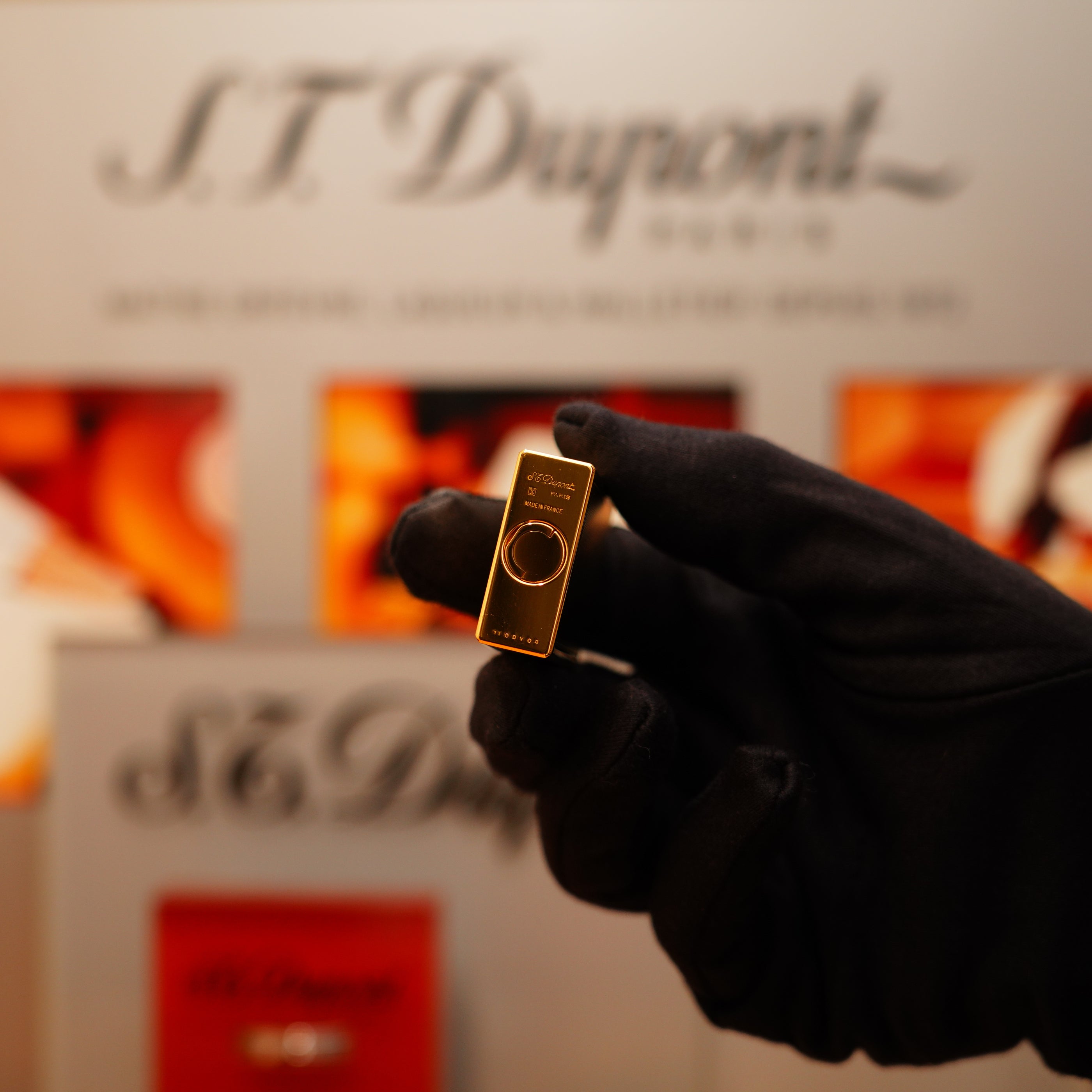 A gloved hand holds a Vintage 1990 S.T. Dupont 24k Large Grain d'Org Iconic Ligne 1 Heavy Finish Lighter, with a blurred logo and branding in the backdrop.