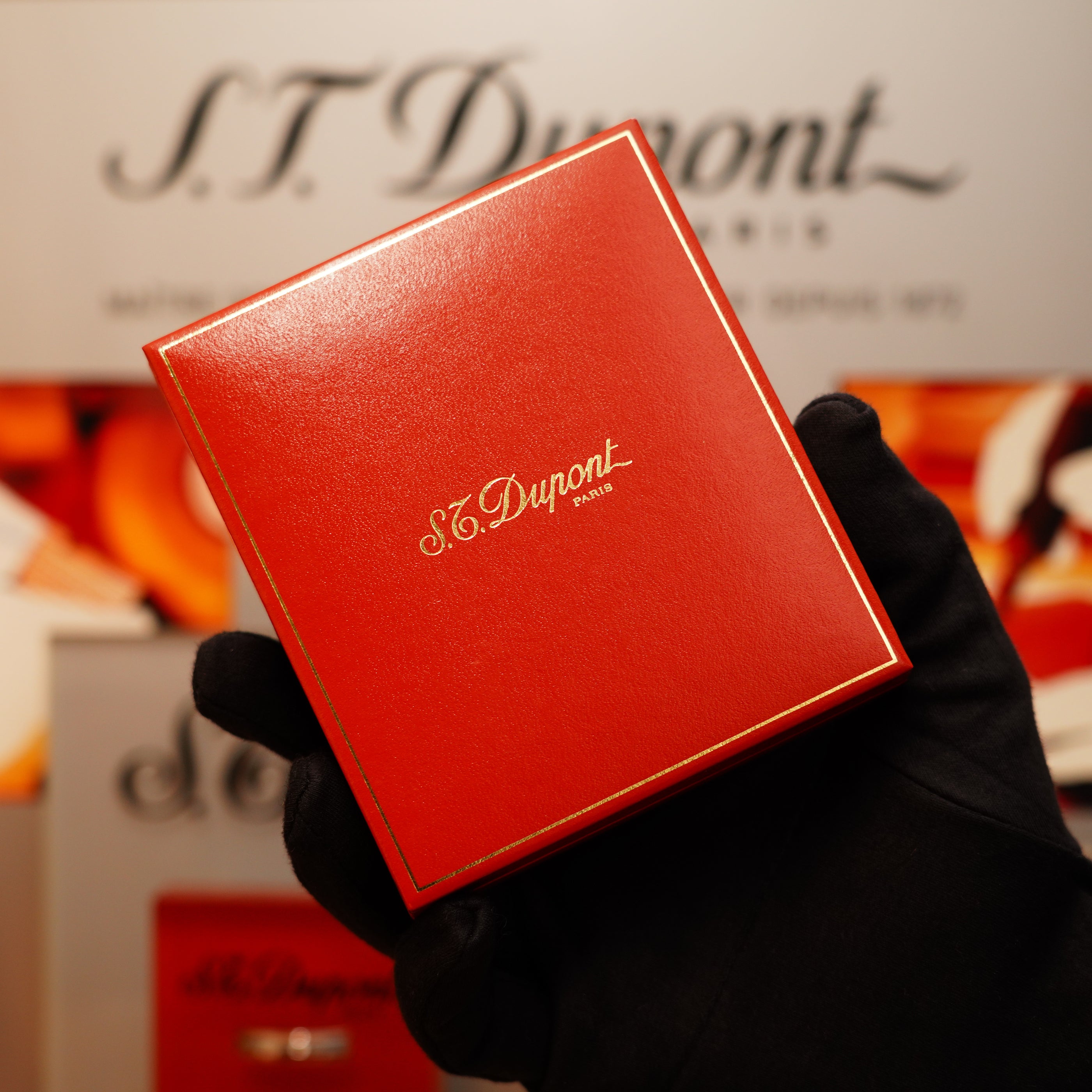 A gloved hand holds a red box containing the Vintage 1990 St Dupont 24k Large Grain d'Org Iconic Ligne 1 Heavy Finish Lighter, with blurred S.T. Dupont branding in the background.