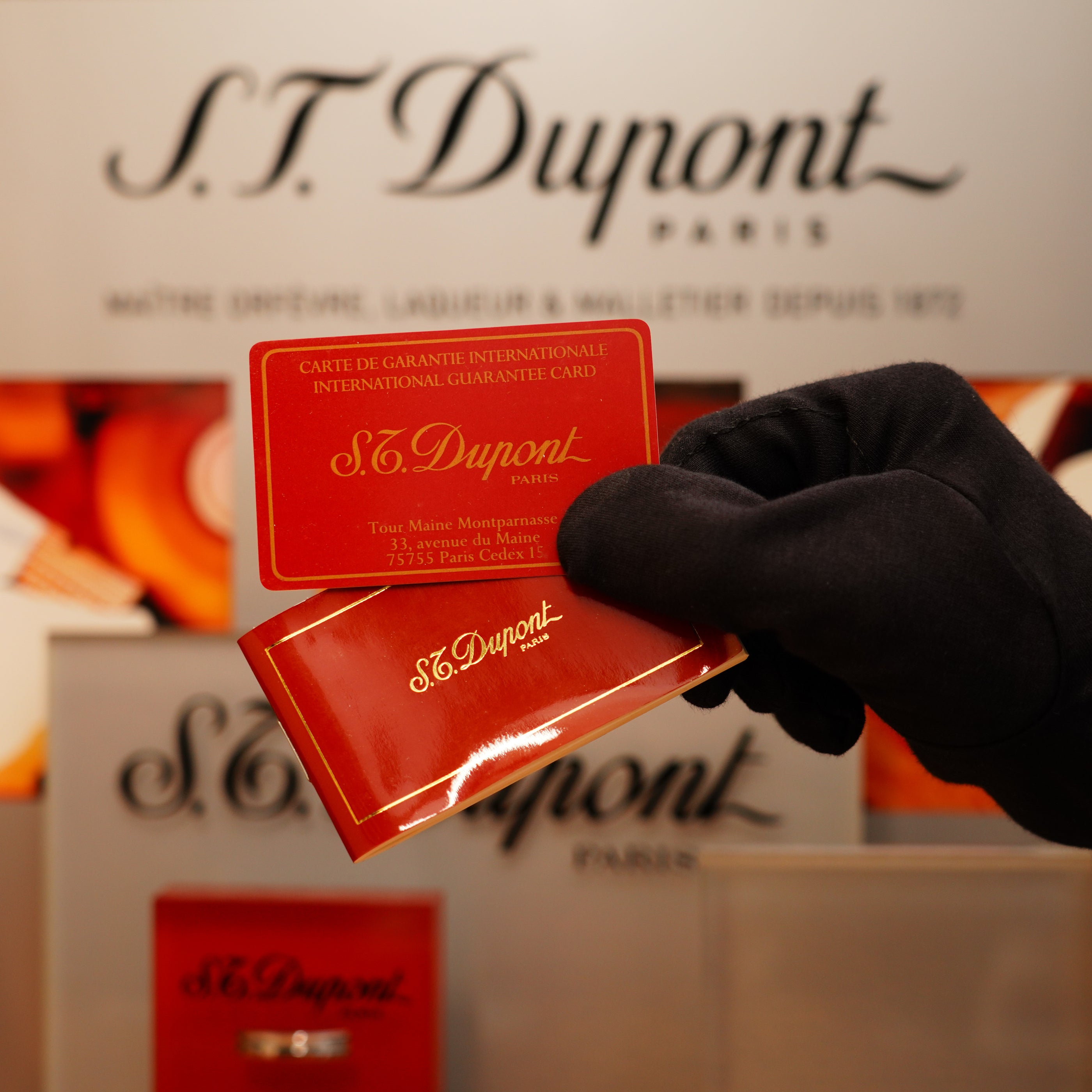 A gloved hand holds an international guarantee card in front of a display featuring the S.T. Dupont logo and orange boxes, showcasing the Vintage 1990 St Dupont 24k Large Grain d'Org Iconic Ligne 1 Heavy Finish Lighter.