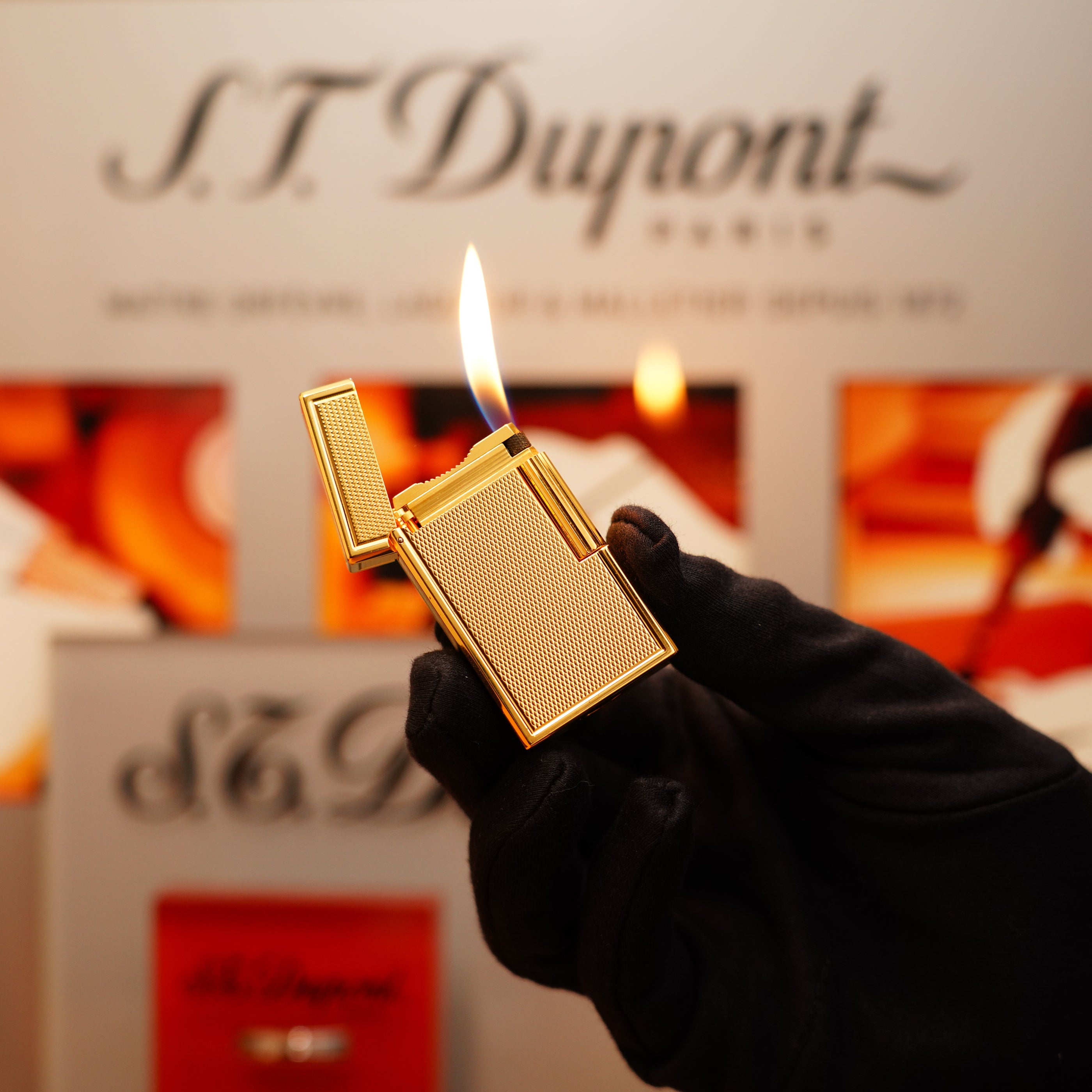 A gloved hand holds a Vintage 1990 S.T. Dupont 24k Large Grain d'Org Iconic Ligne 1 Heavy Finish Lighter, open with a flame, against a blurred S.T. Dupont Paris logo backdrop.