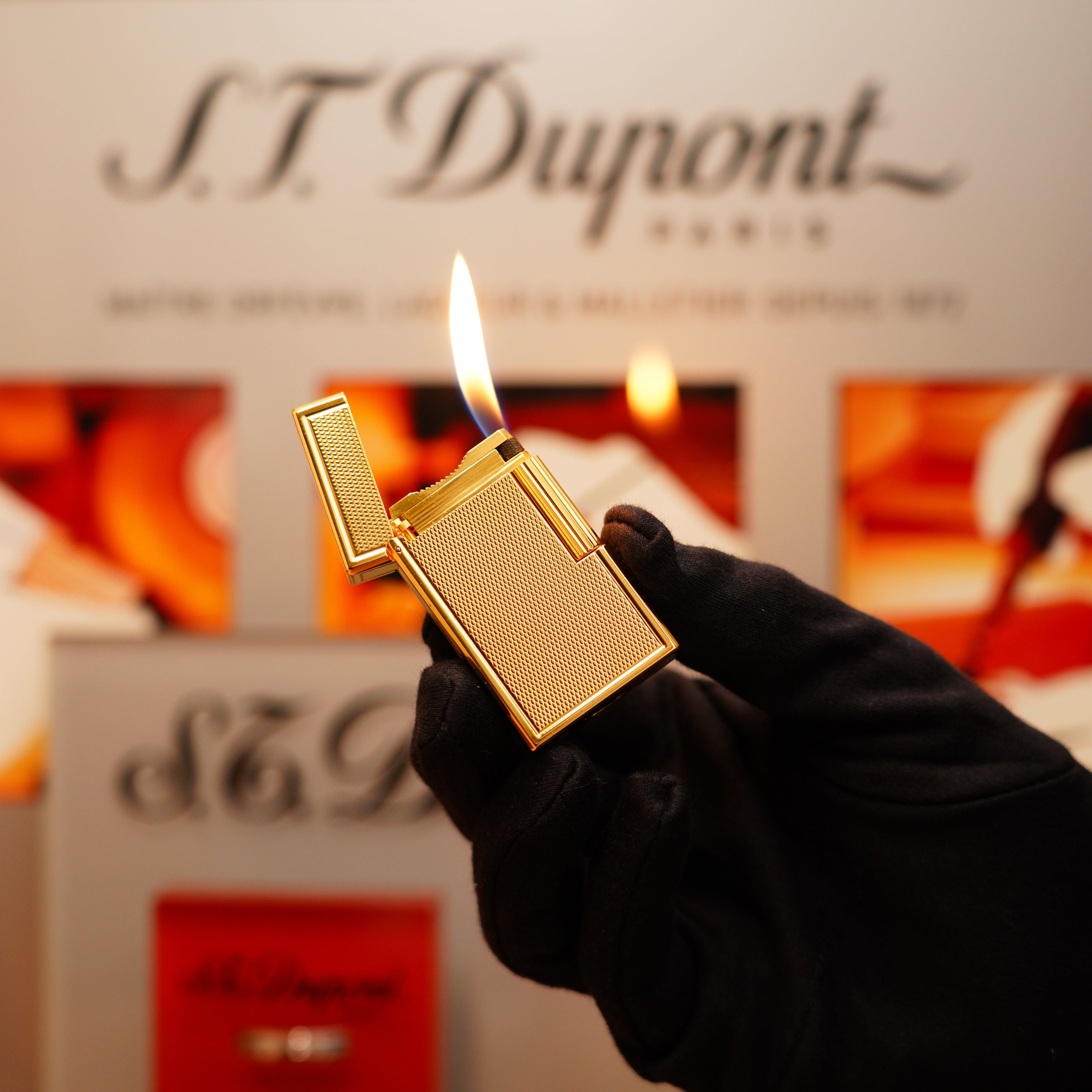 A person in black gloves holds an open Vintage 1990 S.T. Dupont 24k Large Grain d'Org Iconic Ligne 1 Heavy Finish Lighter, its flame flickering against a blurred background showing the brand name "S.T. Dupont.