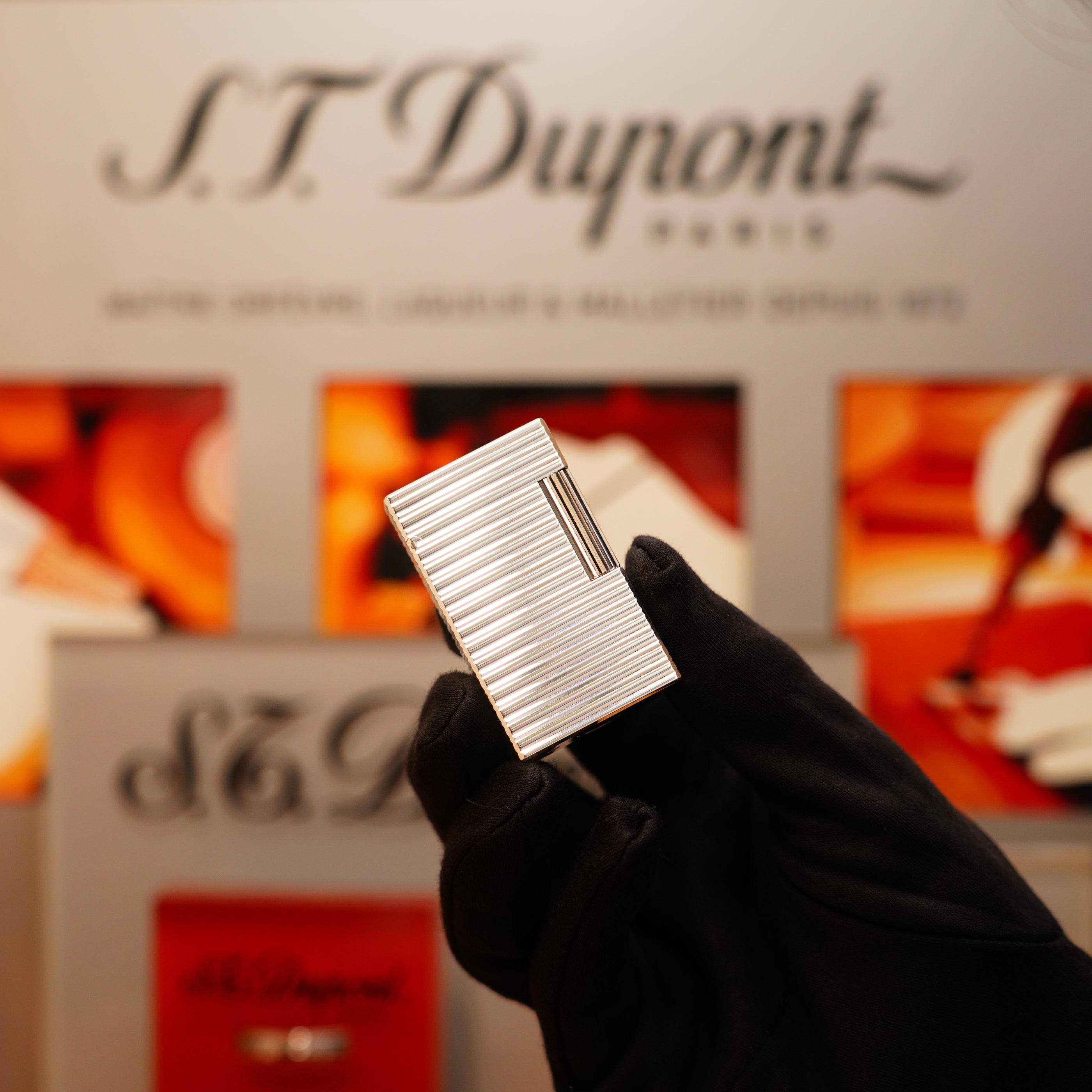 A person wearing black gloves holds a Vintage 1990 S.T. Dupont Large Heavy Silver Finish Ligne 1 Horizontal Pattern Lighter against a backdrop showcasing S.T. Dupont branding.