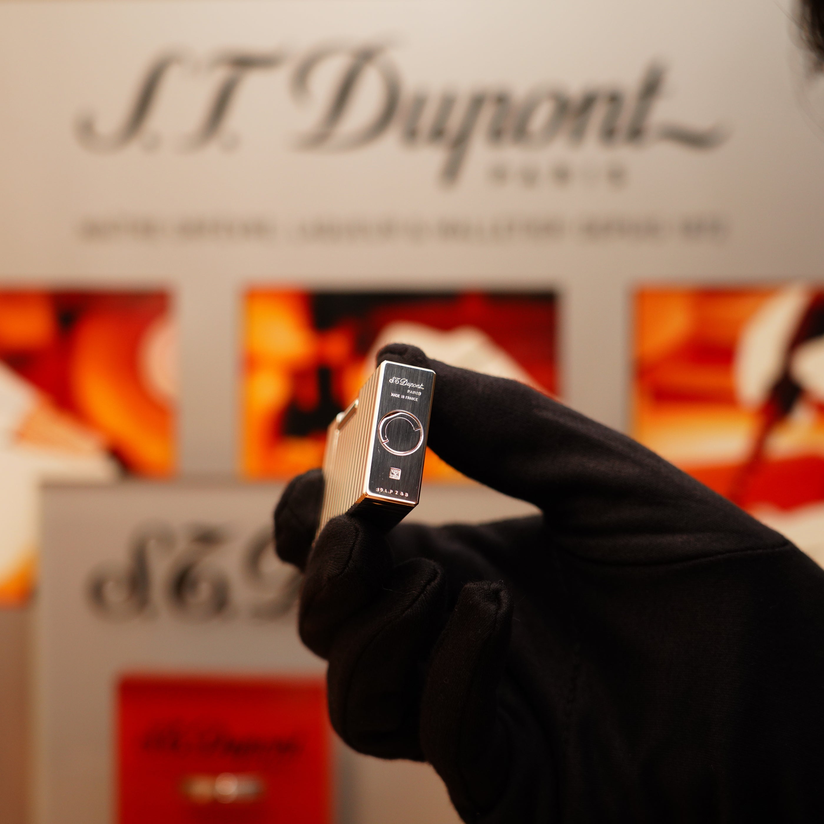 A gloved hand holds a Vintage 1990 S.T. Dupont Ligne 1 Lighter with a heavy silver finish and horizontal pattern against a blurred branded backdrop.