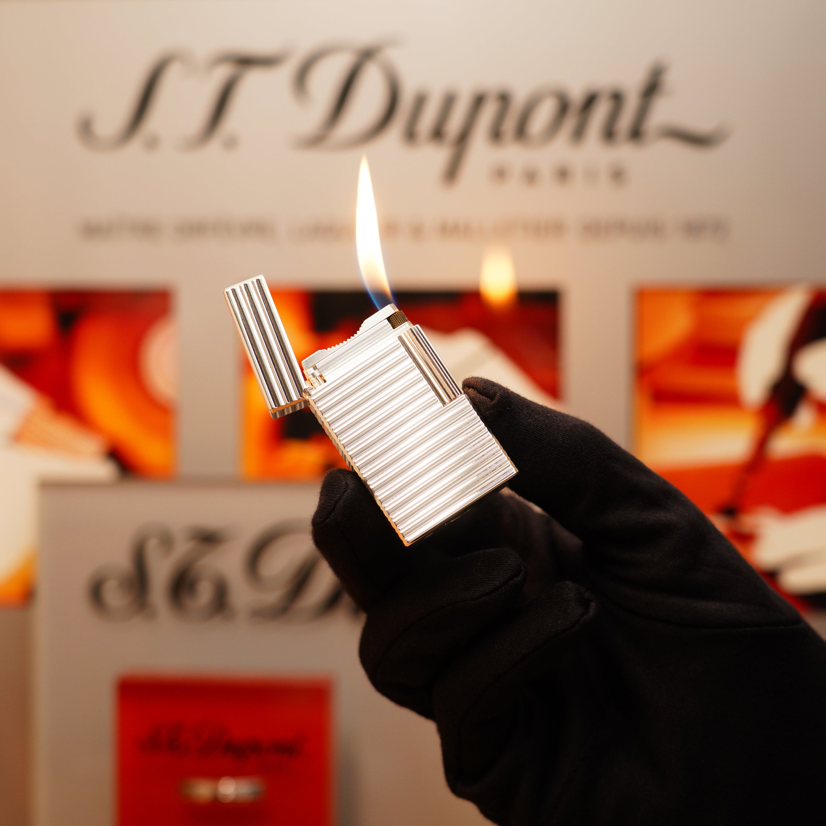 A gloved hand holds a lit S.T. Dupont lighter, specifically the Vintage 1990 Large Heavy Silver Finish Ligne 1 with a Horizontal Pattern, in front of a blurred S.T. Dupont display background.
