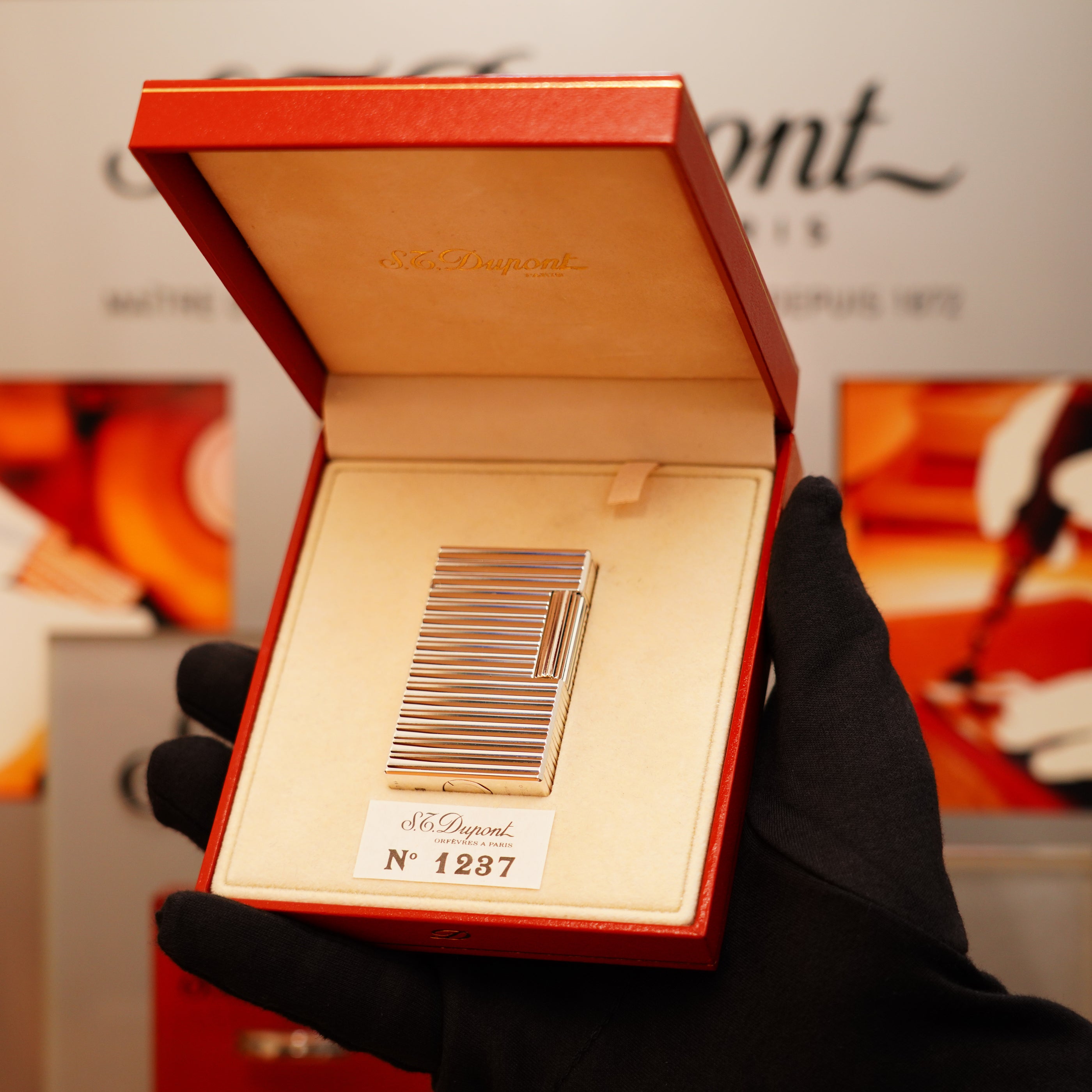 A gloved hand holds an open red box displaying a Vintage 1990 S.T. Dupont large heavy silver finish Ligne 1 horizontal pattern lighter, with "No 1287" against a blurred background featuring the S.T. Dupont logo.