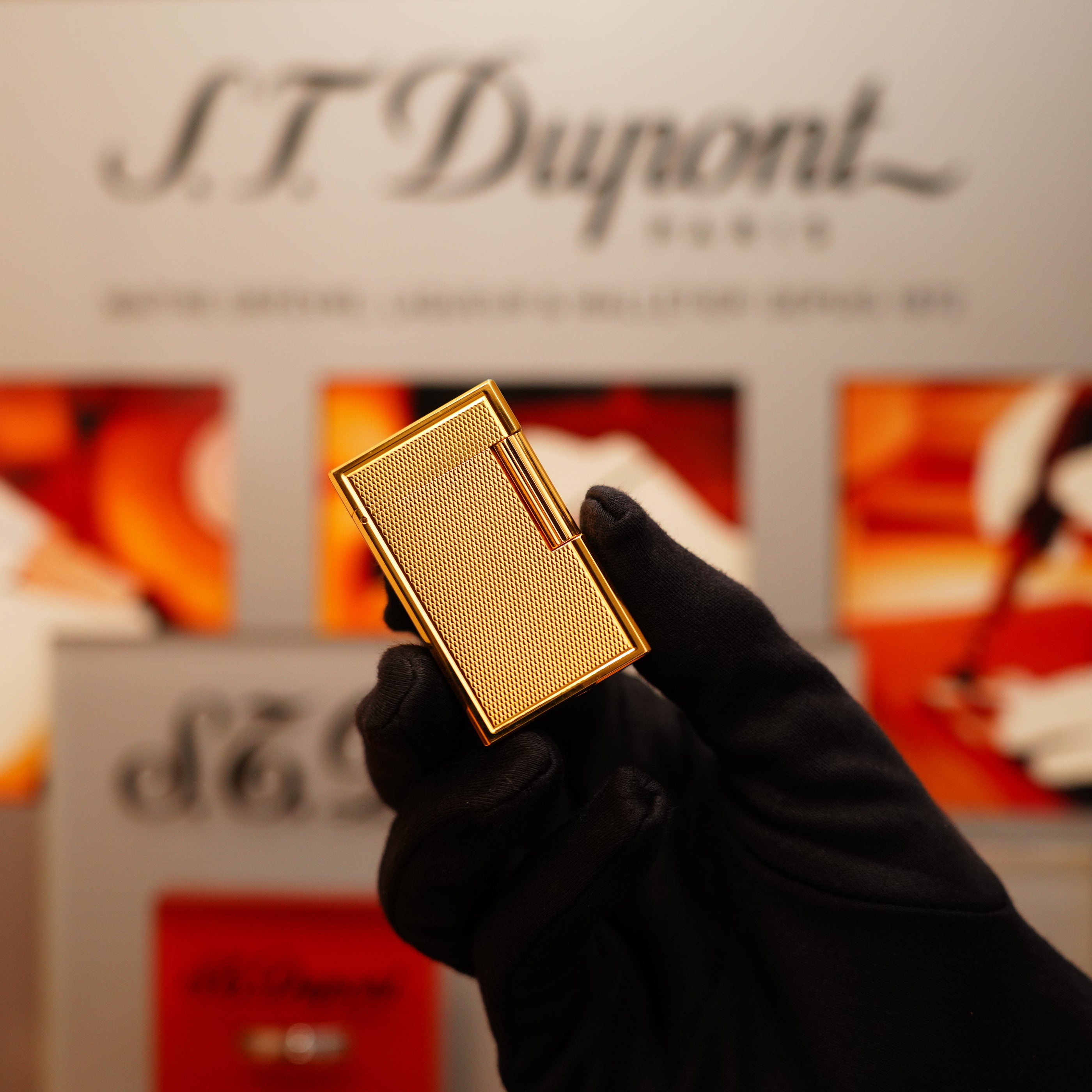 A gloved hand holds a Vintage 1994 S.T. Dupont 24k Large Grain d'Org Iconic Ligne 1 Heavy Finish Lighter against a blurred brand display background.