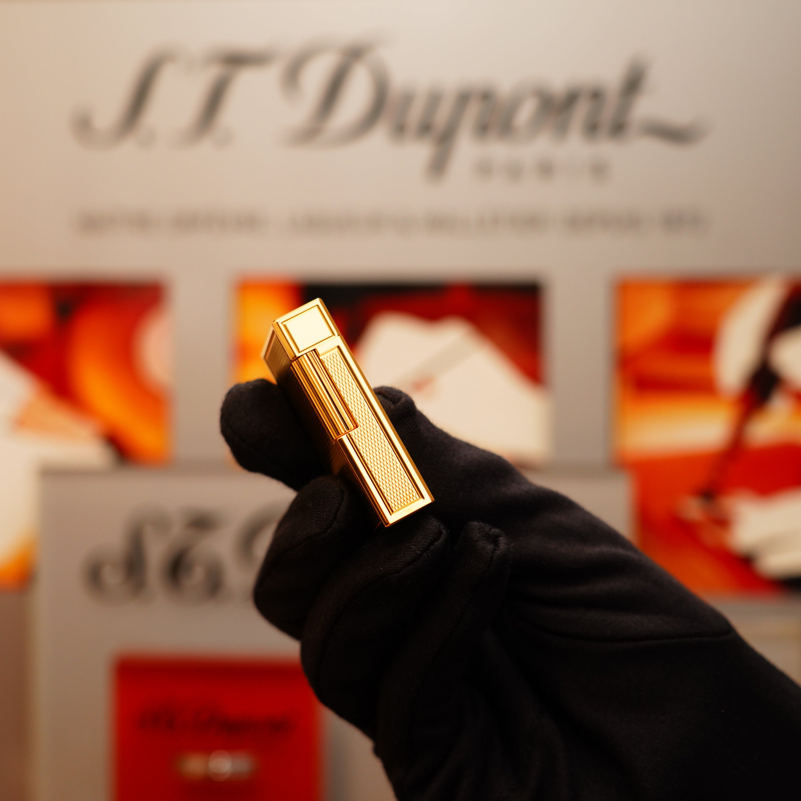 A hand in a black glove holds a Vintage 1994 S.T. Dupont 24k Large Grain d'Org Iconic Ligne 1 Heavy Finish Lighter against a blurred background, featuring red and white elements along with the S.T. Dupont logo.