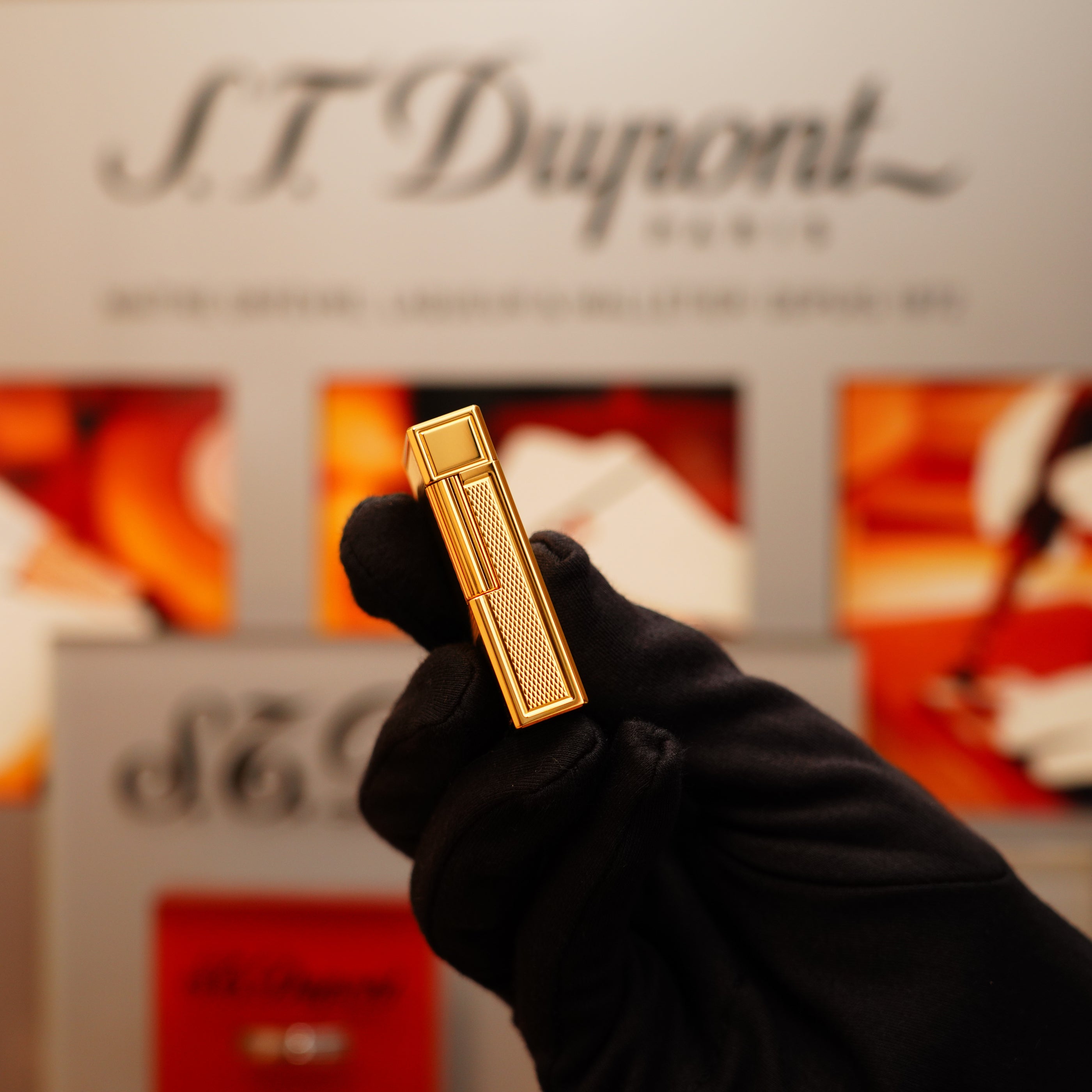 A gloved hand holds a Vintage 1994 St Dupont 24k Large Grain d'Org Iconic Ligne 1 Heavy Finish Lighter against a blurred S.T. Dupont display.