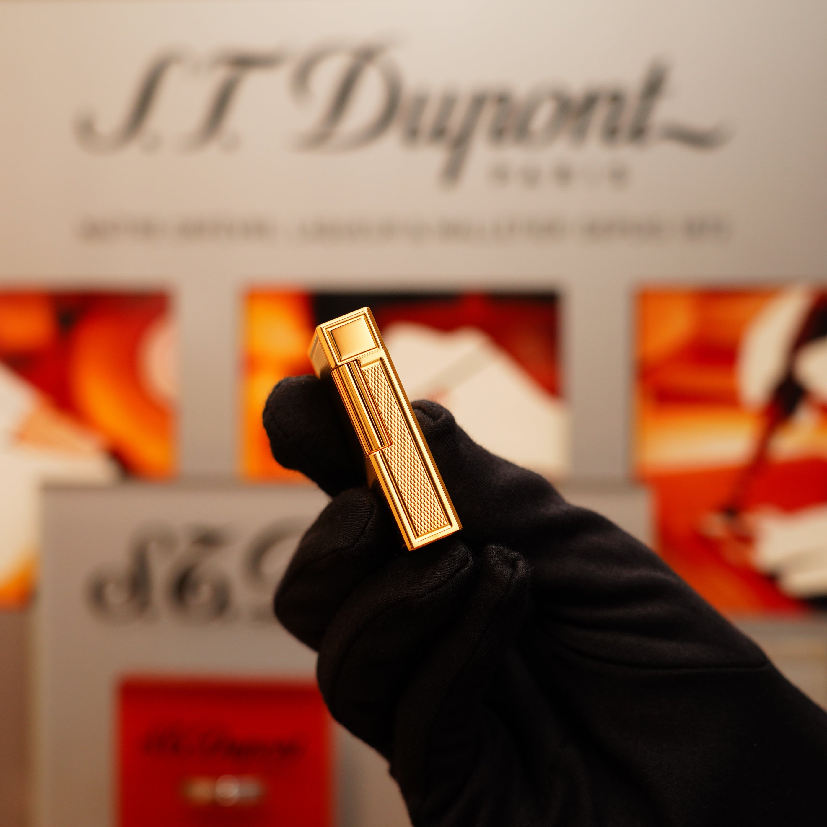 A gloved hand holding a Vintage 1994 S.T. Dupont 24k Large Grain d'Org Iconic Ligne 1 Heavy Finish Lighter with a blurred branded backdrop.
