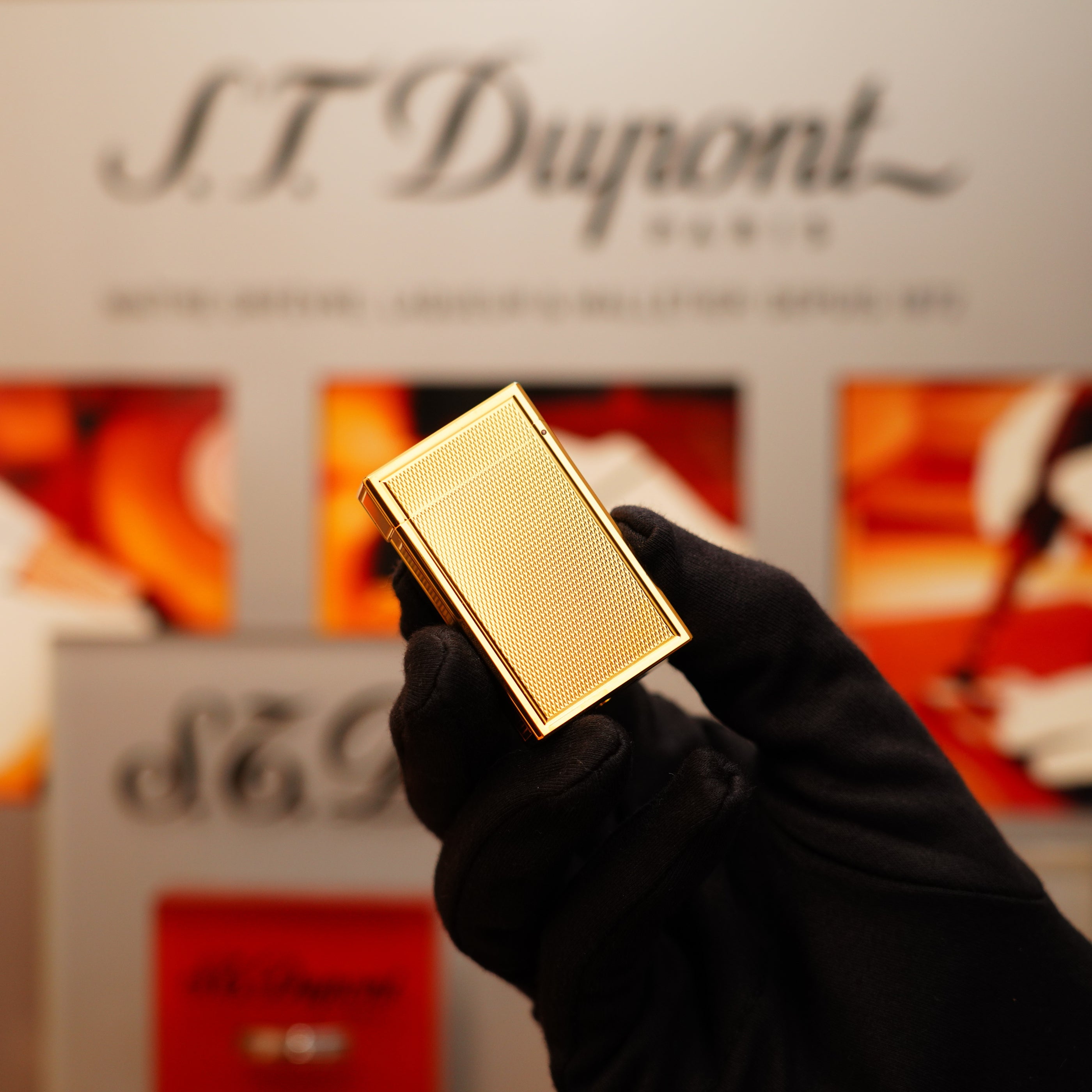 A gloved hand holds a Vintage 1994 St Dupont 24k Large Grain d'Org Iconic Ligne 1 heavy finish lighter against a blurred background featuring S.T. Dupont branding.