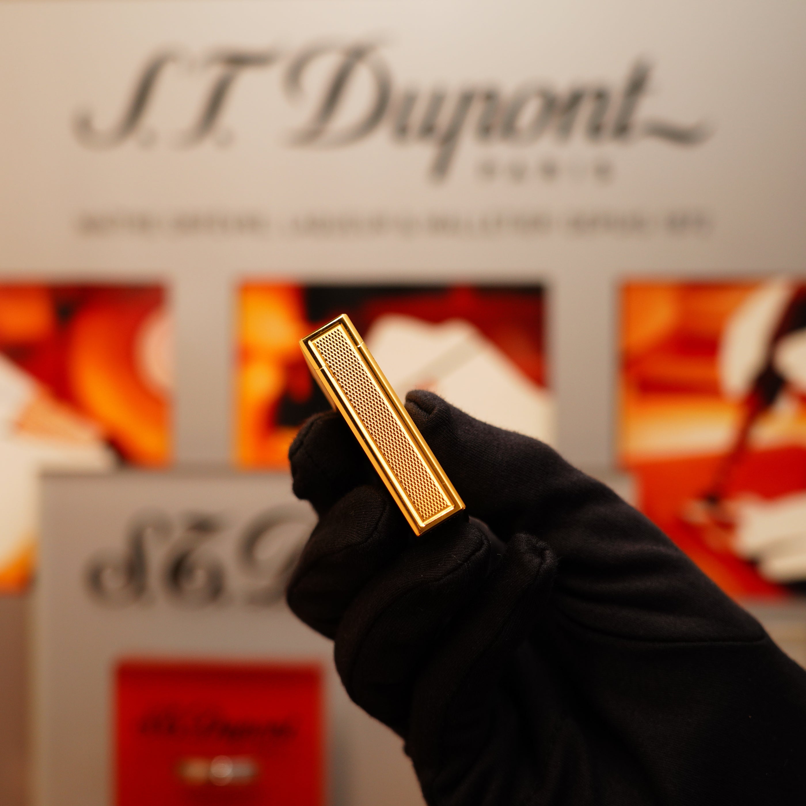 A gloved hand holds a Vintage 1994 S.T. Dupont 24k Large Grain d'Org Iconic Ligne 1 Heavy Finish Lighter in front of a blurred S.T. Dupont display.