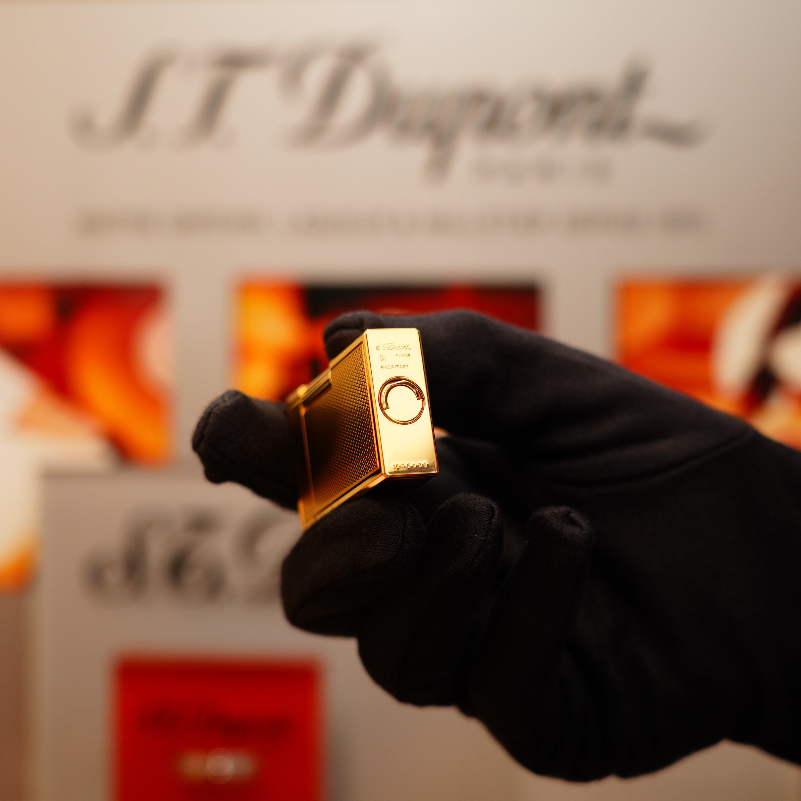 A gloved hand holds a Vintage 1994 St Dupont 24k Large Grain d'Org Iconic Ligne 1 Heavy Finish Lighter in front of a blurred background displaying the S.T. Dupont logo.