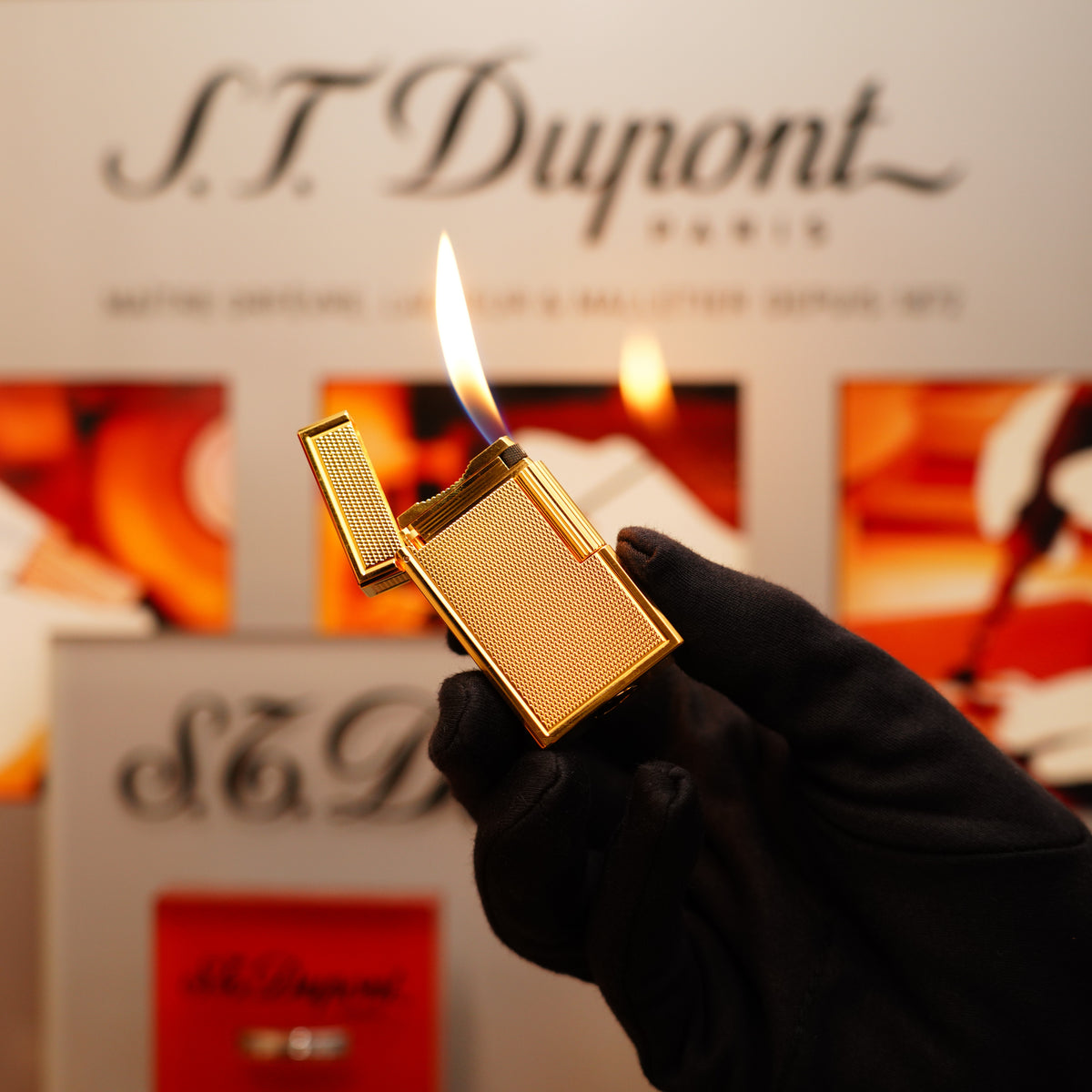 A gloved hand holds a Vintage 1994 St Dupont 24k Large Grain d'Org Iconic Ligne 1 lighter with a flame against a blurred background showcasing S.T. Dupont branding.