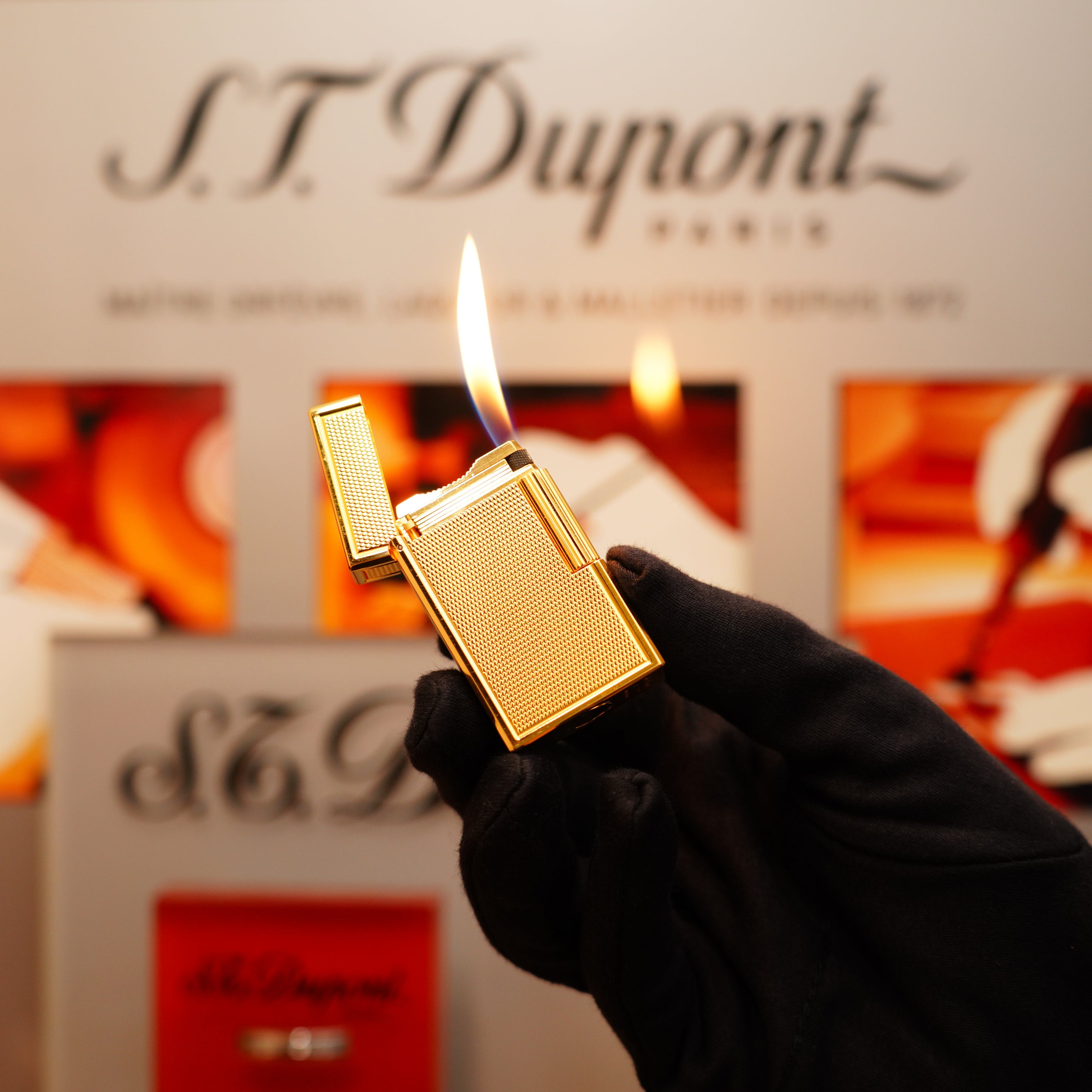A gloved hand holds a lit Vintage 1994 S.T. Dupont 24k Large Grain d'Org Iconic Ligne 1 Heavy Finish Lighter against a blurred backdrop featuring the S.T. Dupont logo.