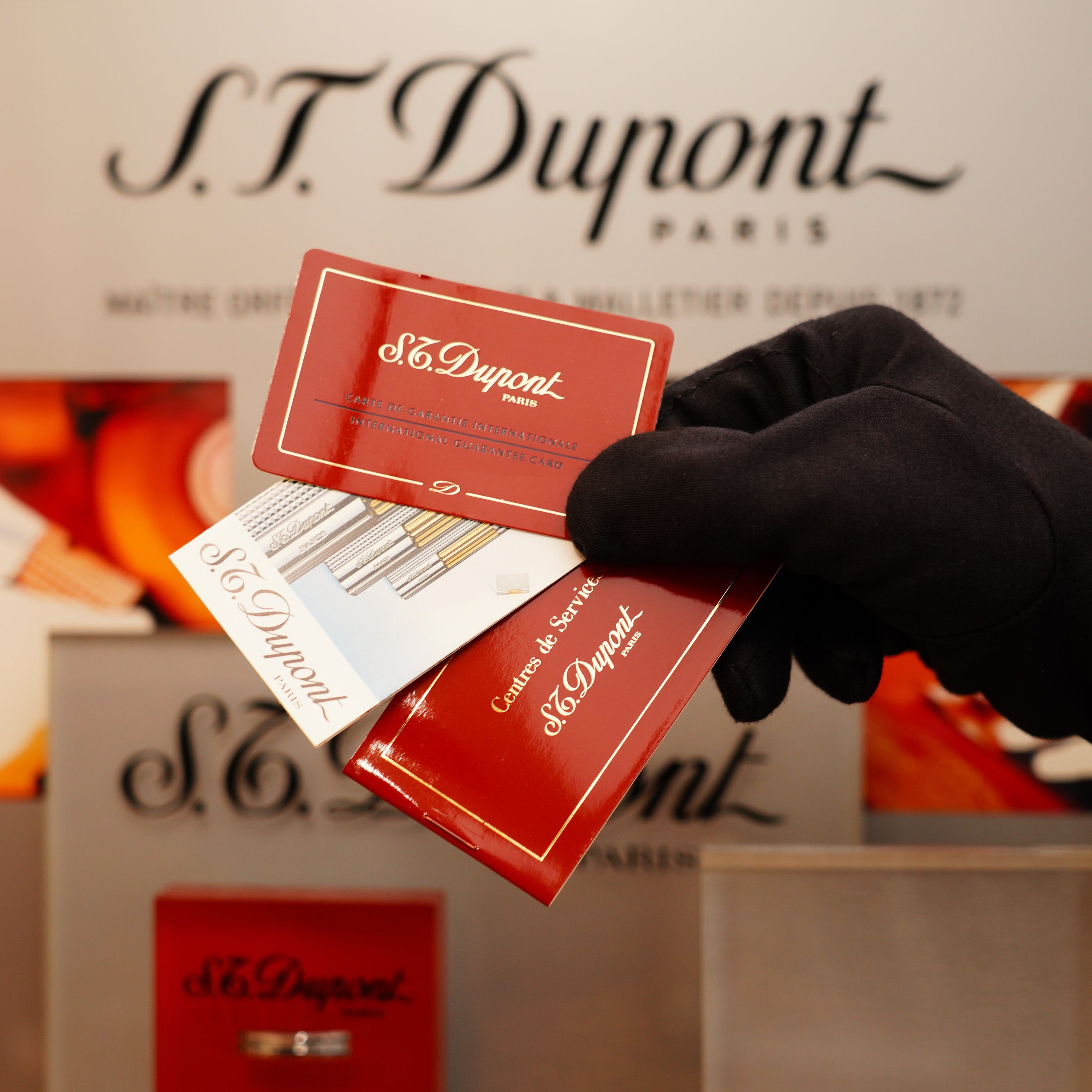 A gloved hand holds a red S.T. Dupont card and booklet in front of an S.T. Dupont Paris display, featuring the Vintage 1994 Ligne 1 Heavy Finish Lighter with 24k gold and large grain d'org design.