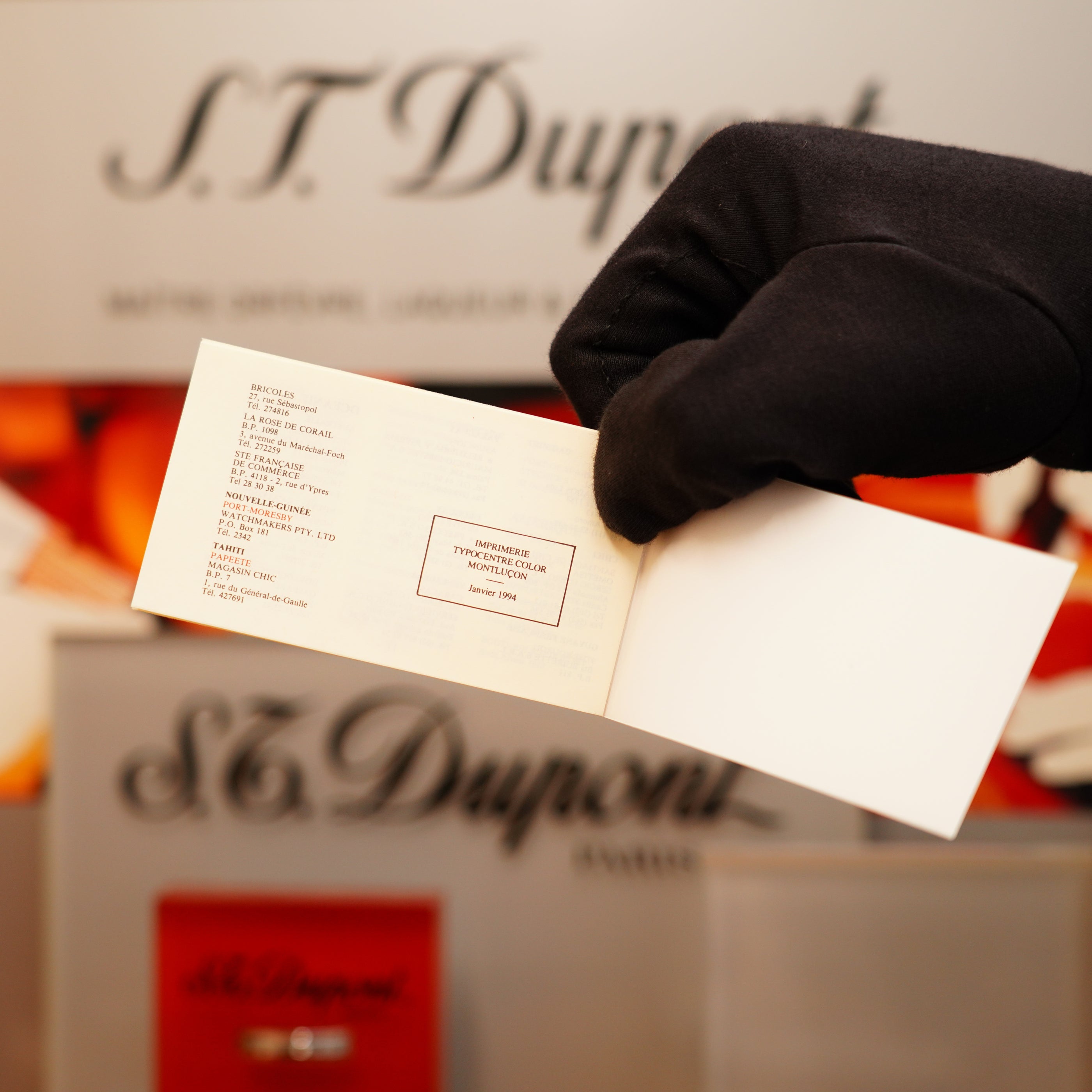 A gloved hand holds open a small booklet with text details in front of a display featuring the Vintage 1994 S.T. Dupont 24k Large Grain d'Org Iconic Ligne 1 Heavy Finish Lighter.