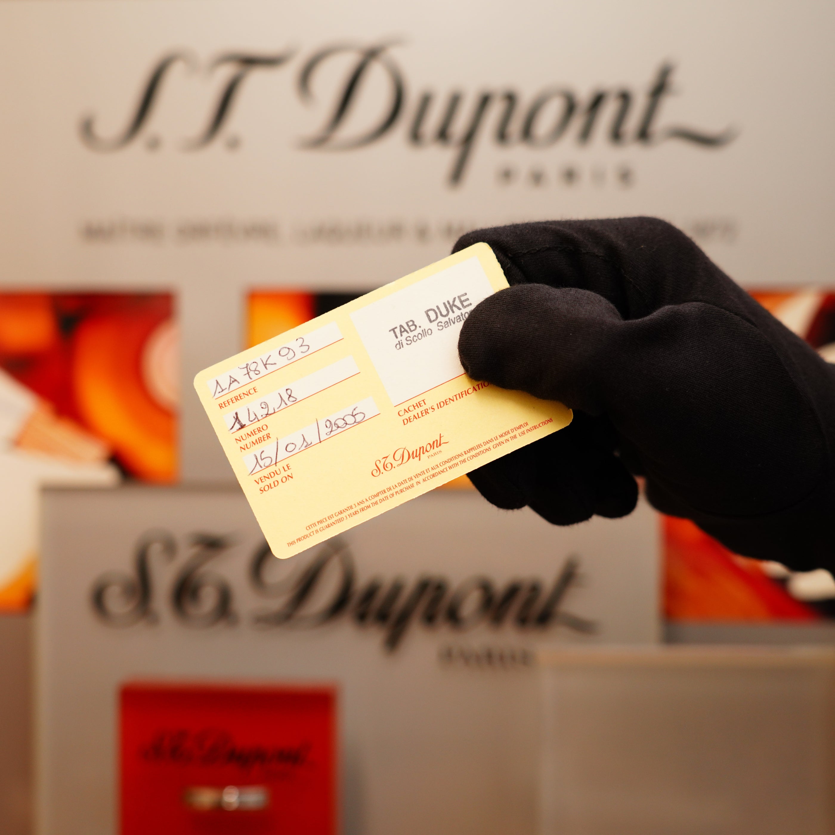 A gloved hand holds a small yellow card in front of a display featuring the S.T. Dupont logo, showcasing the Vintage 1994 St Dupont 24k Large Grain d'Org Iconic Ligne 1 Heavy Finish Lighter.