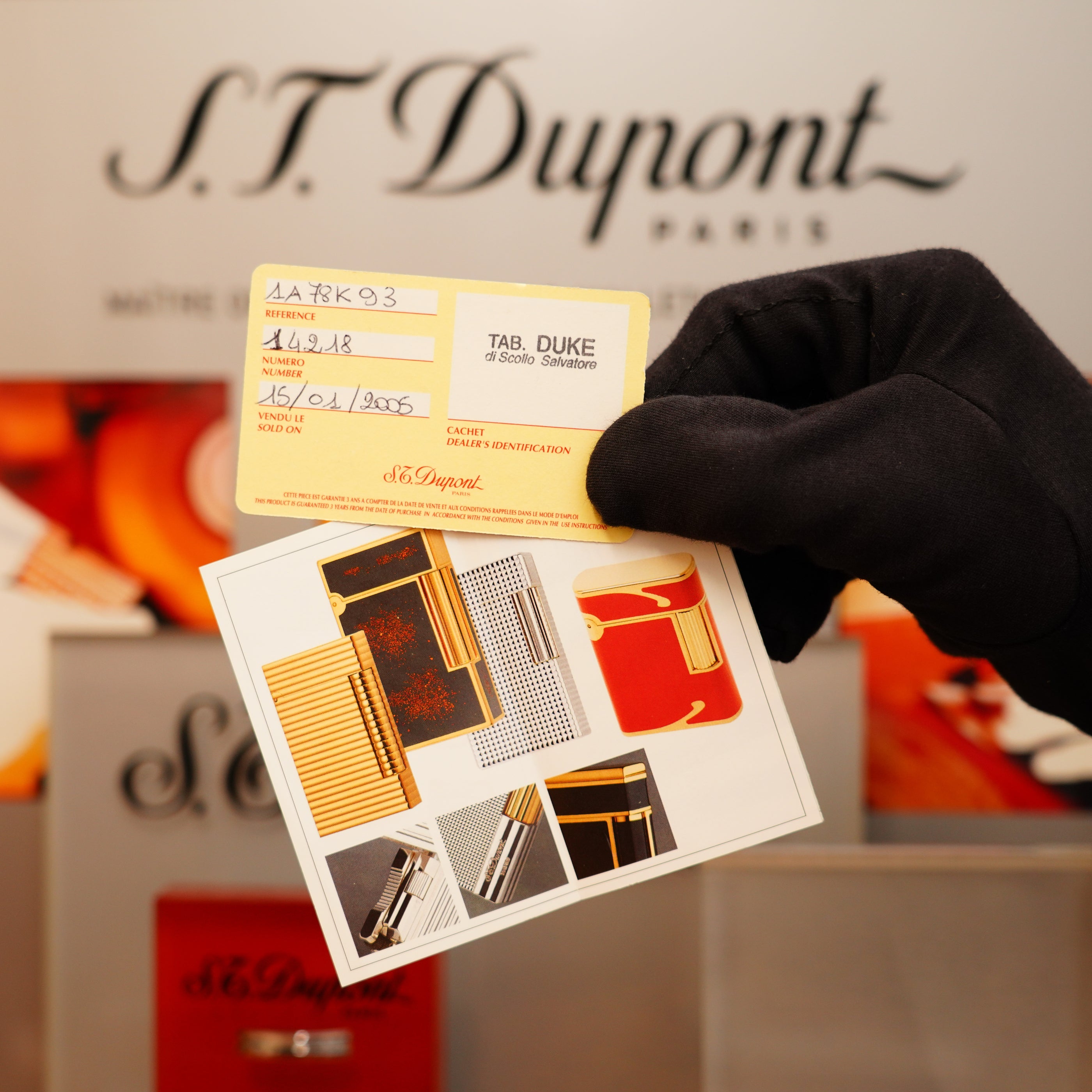 A gloved hand holds a product warranty card and an image card in front of a display featuring the Vintage 1994 S.T. Dupont 24k Large Grain d'Org Iconic Ligne 1 Heavy Finish Lighter.