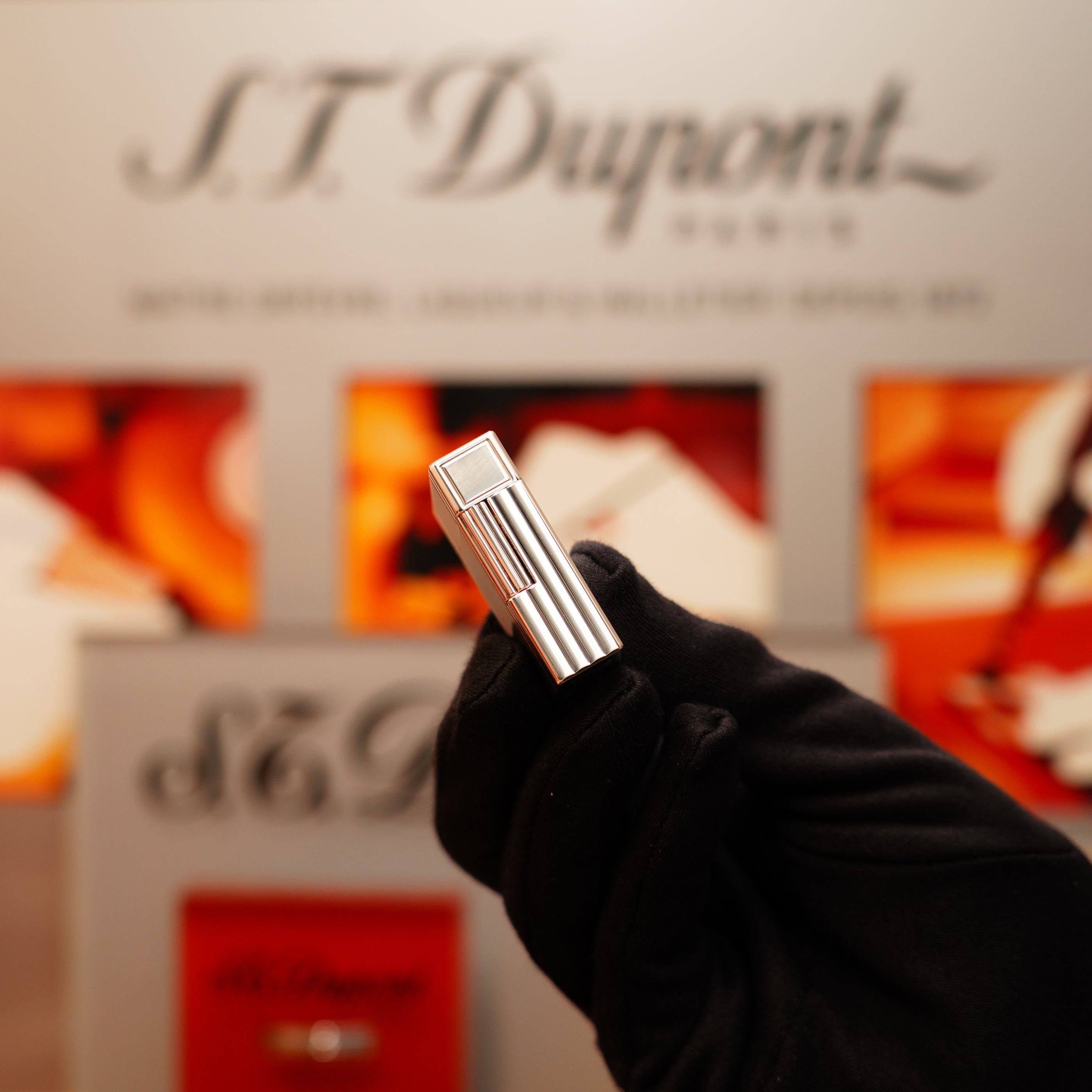 A gloved hand presents a Vintage 2000 S.T. Dupont Heavy Silver Finish Small Ligne 1 Vertical Ligne Lighter against a blurred S.T. Dupont logo backdrop.