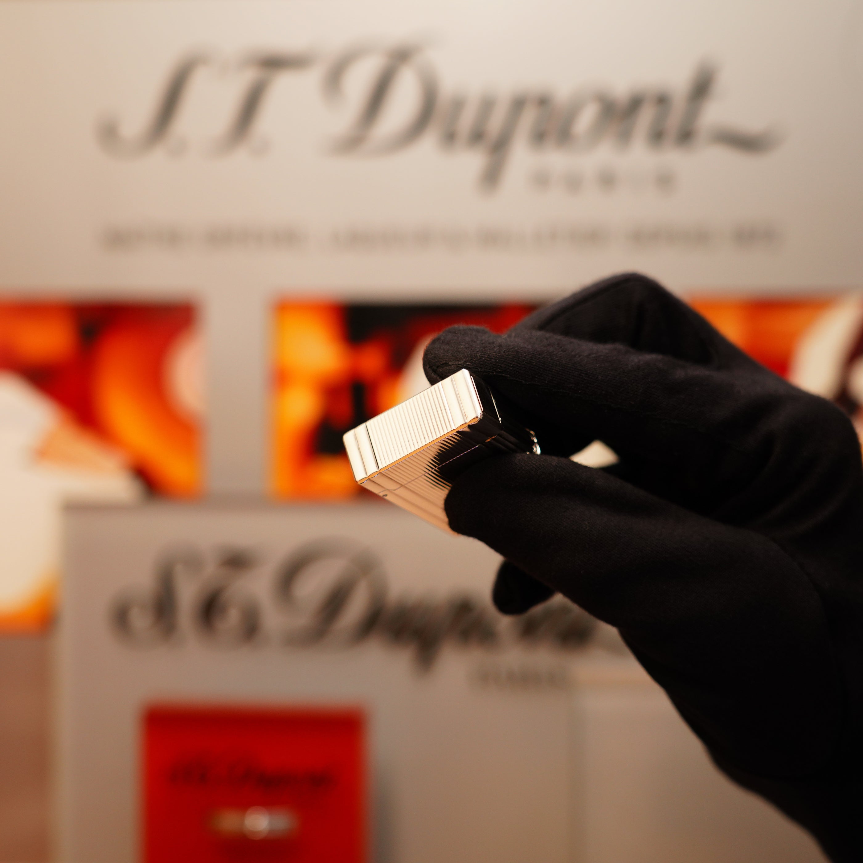 A gloved hand holds a Vintage 2000 S.T. Dupont Heavy Silver Finish Small Ligne 1 Vertical Ligne Lighter, set against a blurred S.T. Dupont display background.