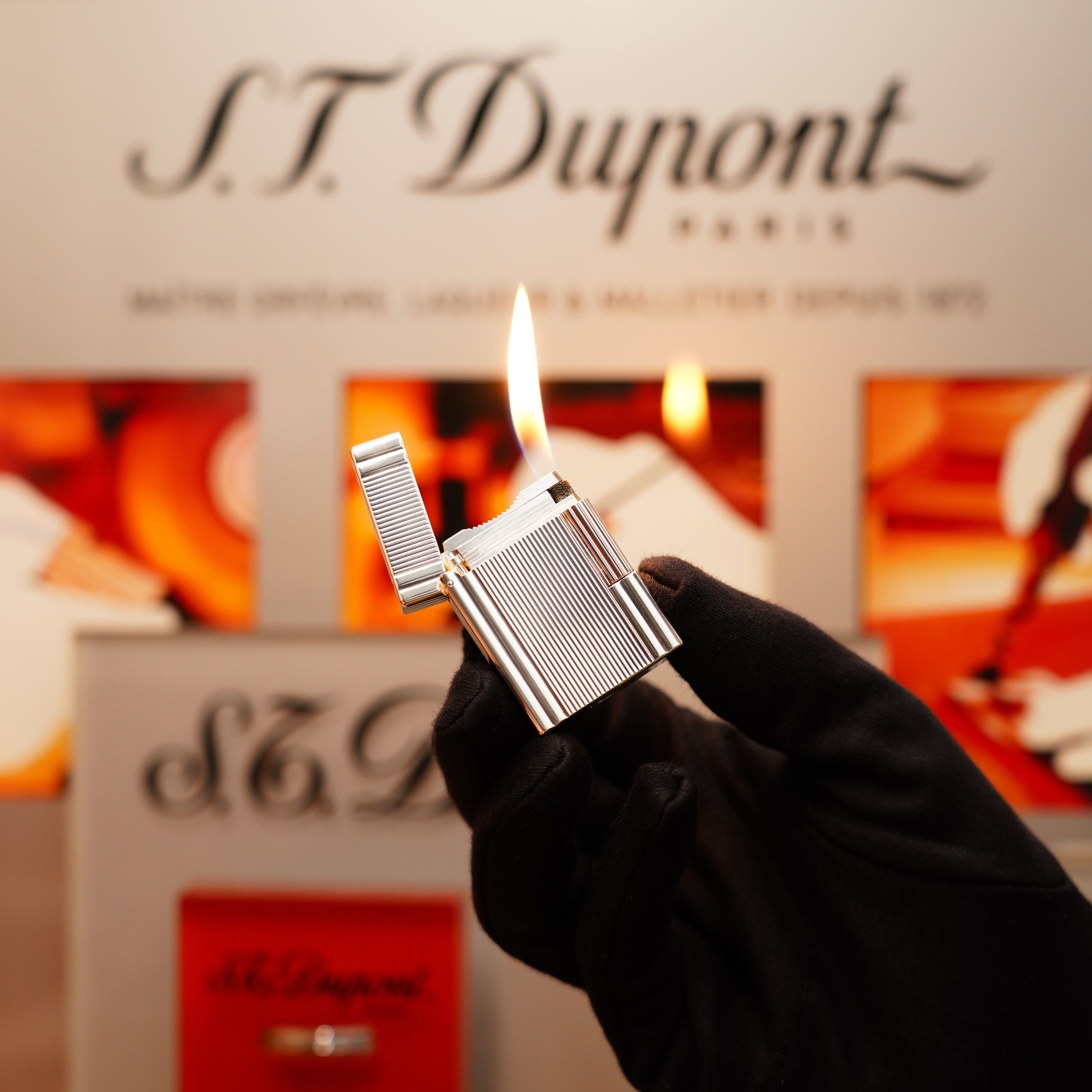 A gloved hand holds a lit Vintage 2000 St Dupont Heavy Silver Finish Small iconic Ligne 1 Vertical Ligne Lighter against a blurred background featuring S.T. Dupont branding.