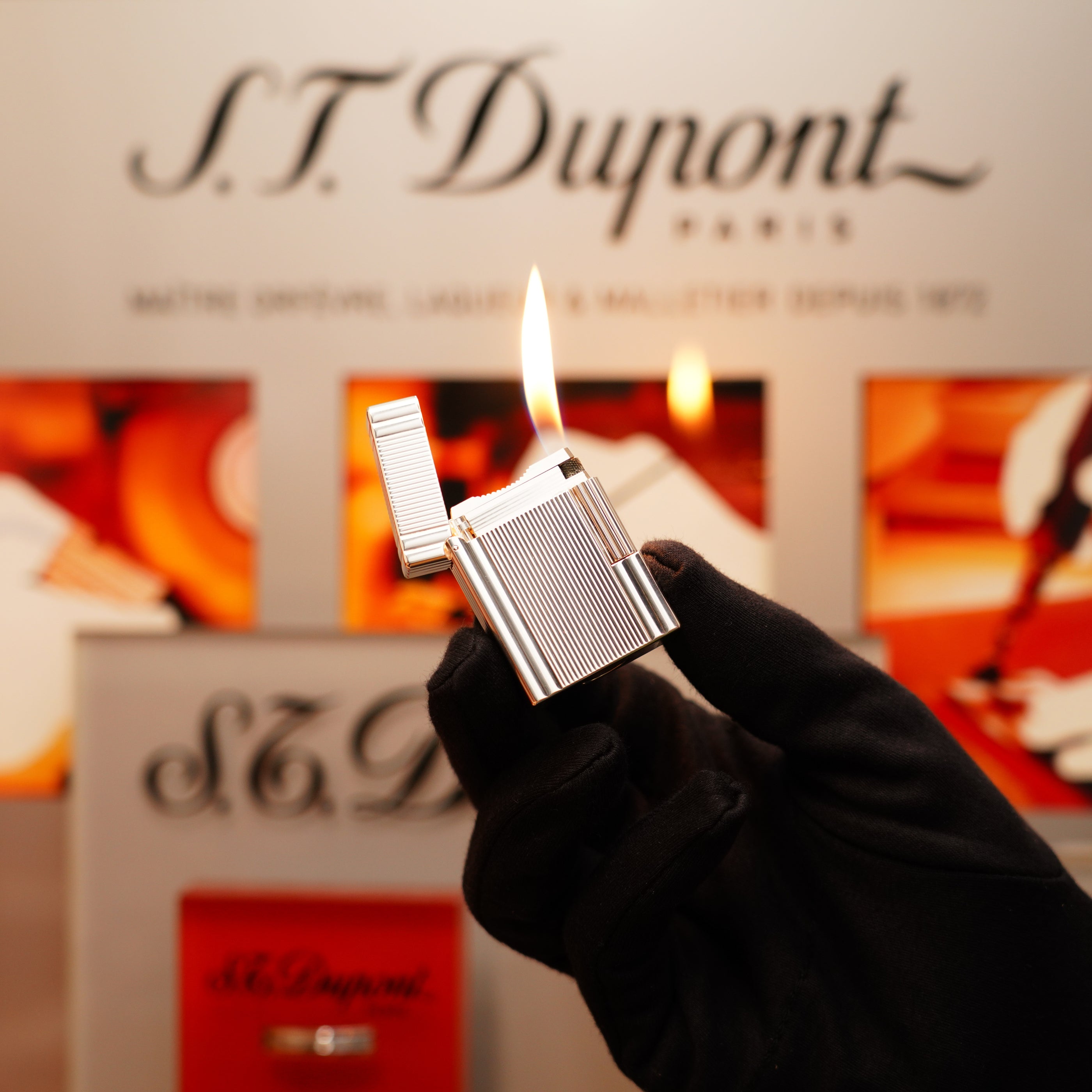 A gloved hand holds a lit Vintage 2000 St Dupont Heavy Silver Finish Small iconic Ligne 1 Vertical Ligne Lighter against a blurred background with the S.T. Dupont logo.