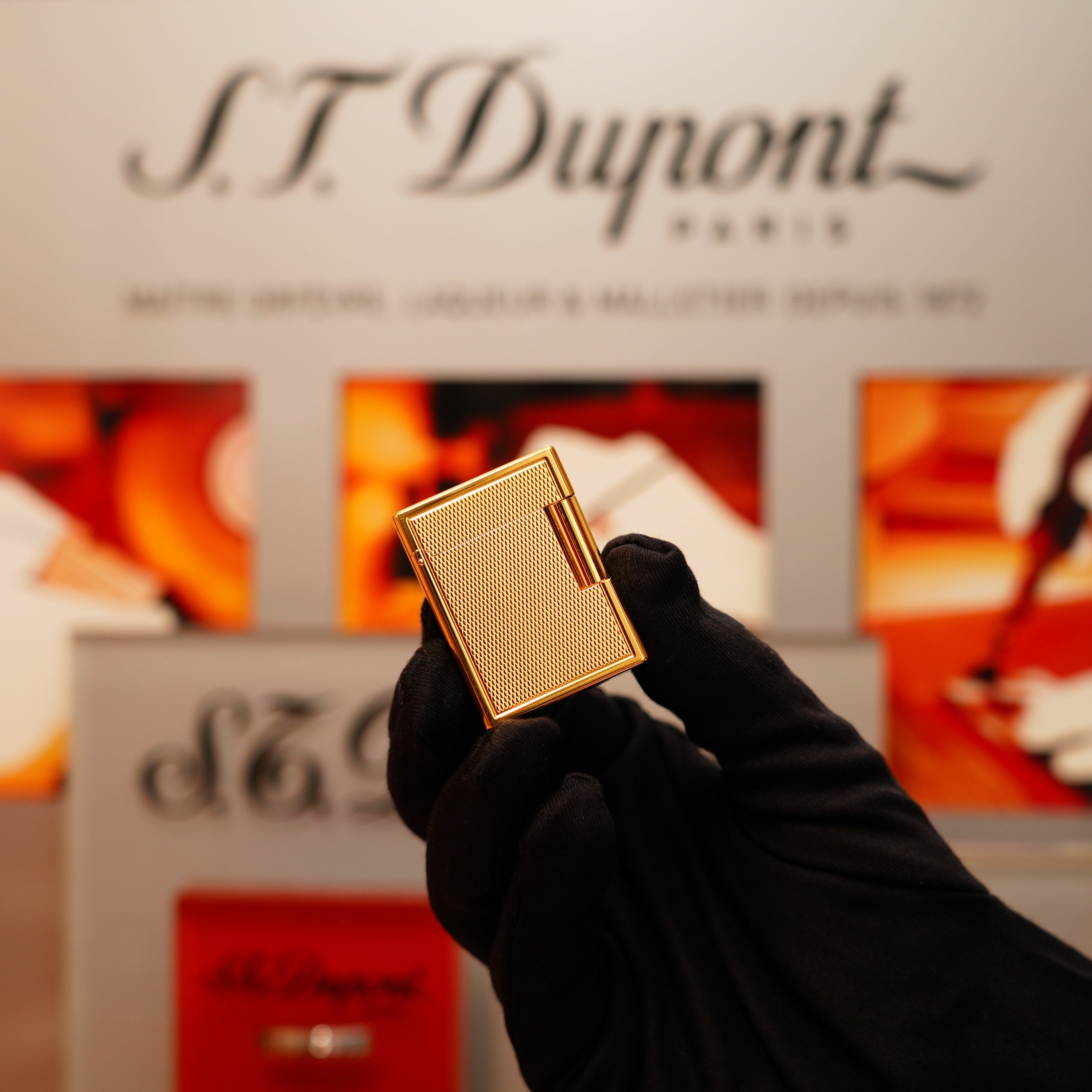 A gloved hand holds a Vintage 1996 S.T. Dupont 24k Small Grain d'Org Iconic Ligne 1 Heavy Finish lighter in front of a blurred S.T. Dupont Paris display.