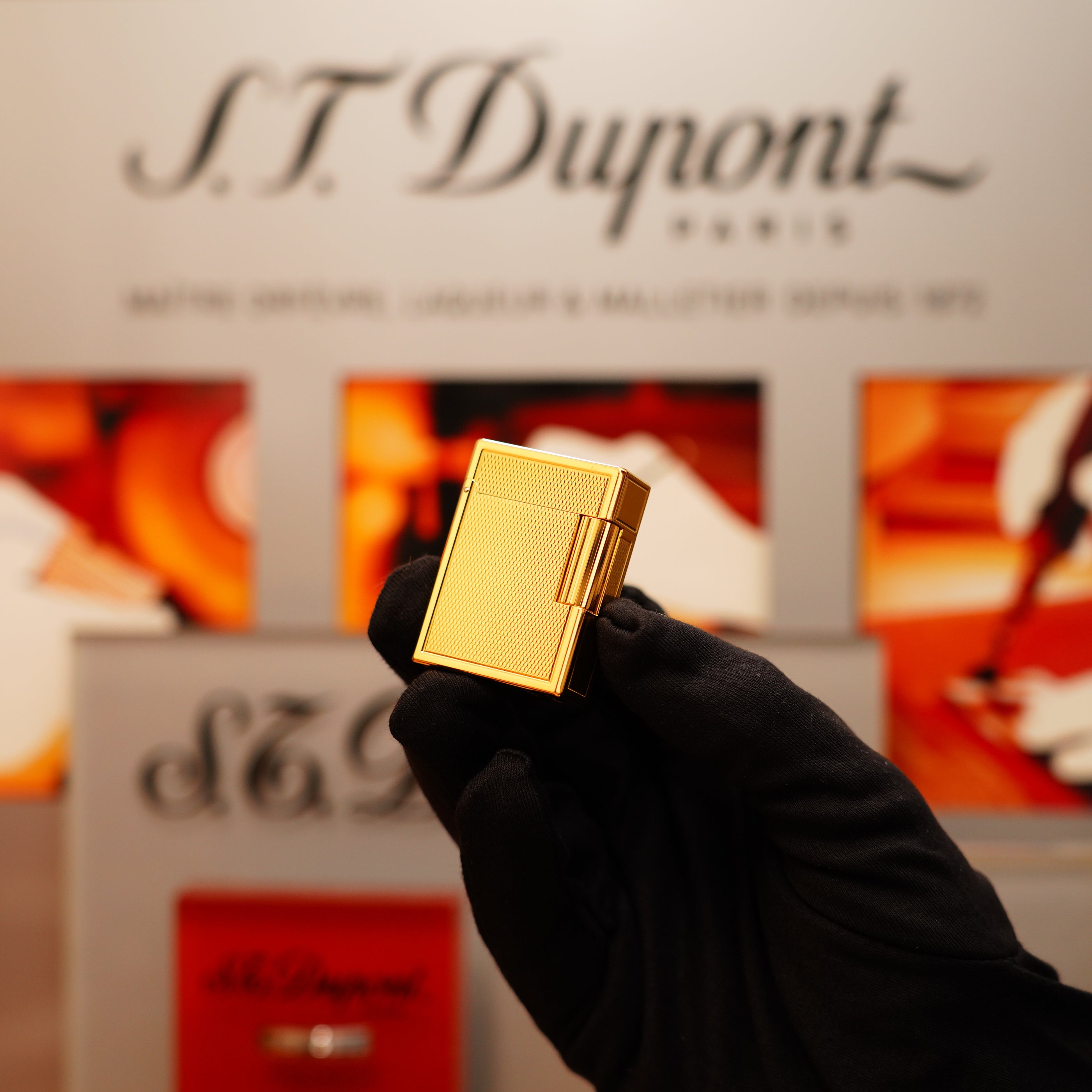 A gloved hand holds a Vintage 1996 St Dupont 24k Small Grain d'Org Iconic Ligne 1 Heavy Finish Lighter, with a blurred, branded background.
