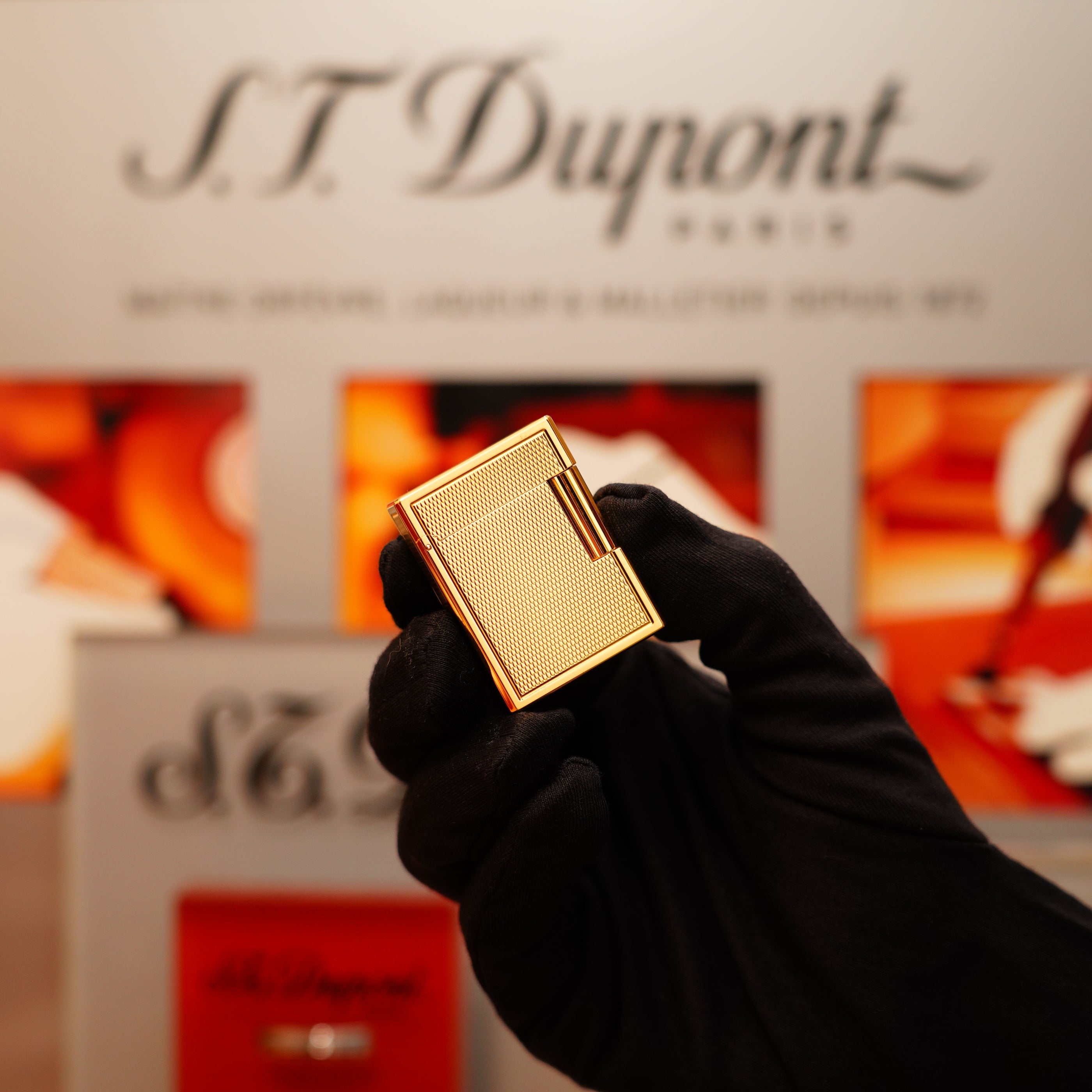 A gloved hand holds a Vintage 1996 S.T. Dupont 24k Small Grain d'Org Iconic Ligne 1 Heavy Finish Lighter against a blurred background featuring the brand's logo and various product images.
