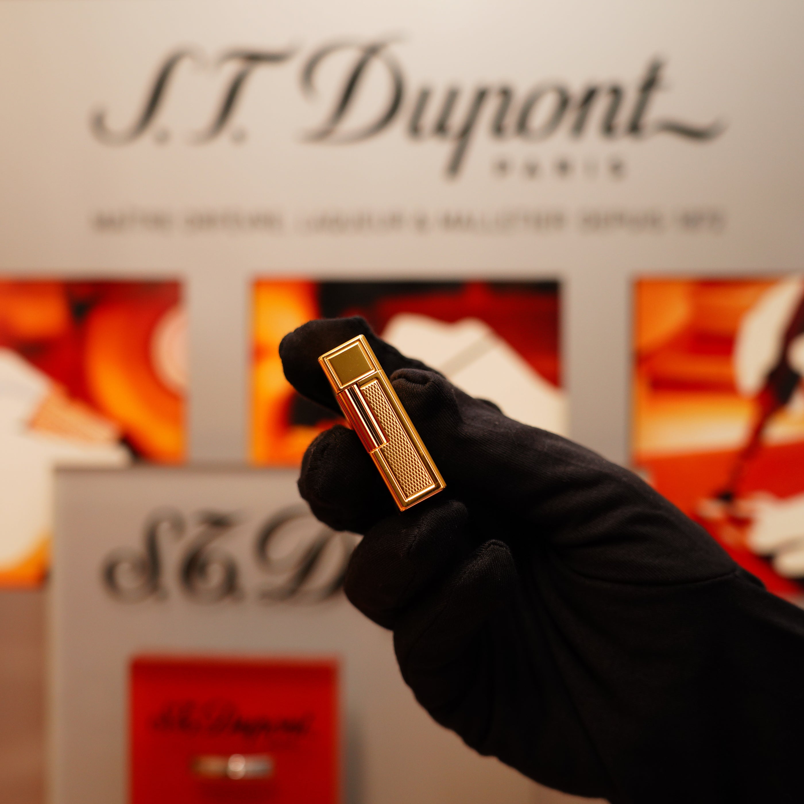 A gloved hand holds a gold Vintage 1996 S.T. Dupont 24k Small Grain d'Org Ligne 1 lighter against a blurred S.T. Dupont logo background in vibrant red, white, and orange tones.