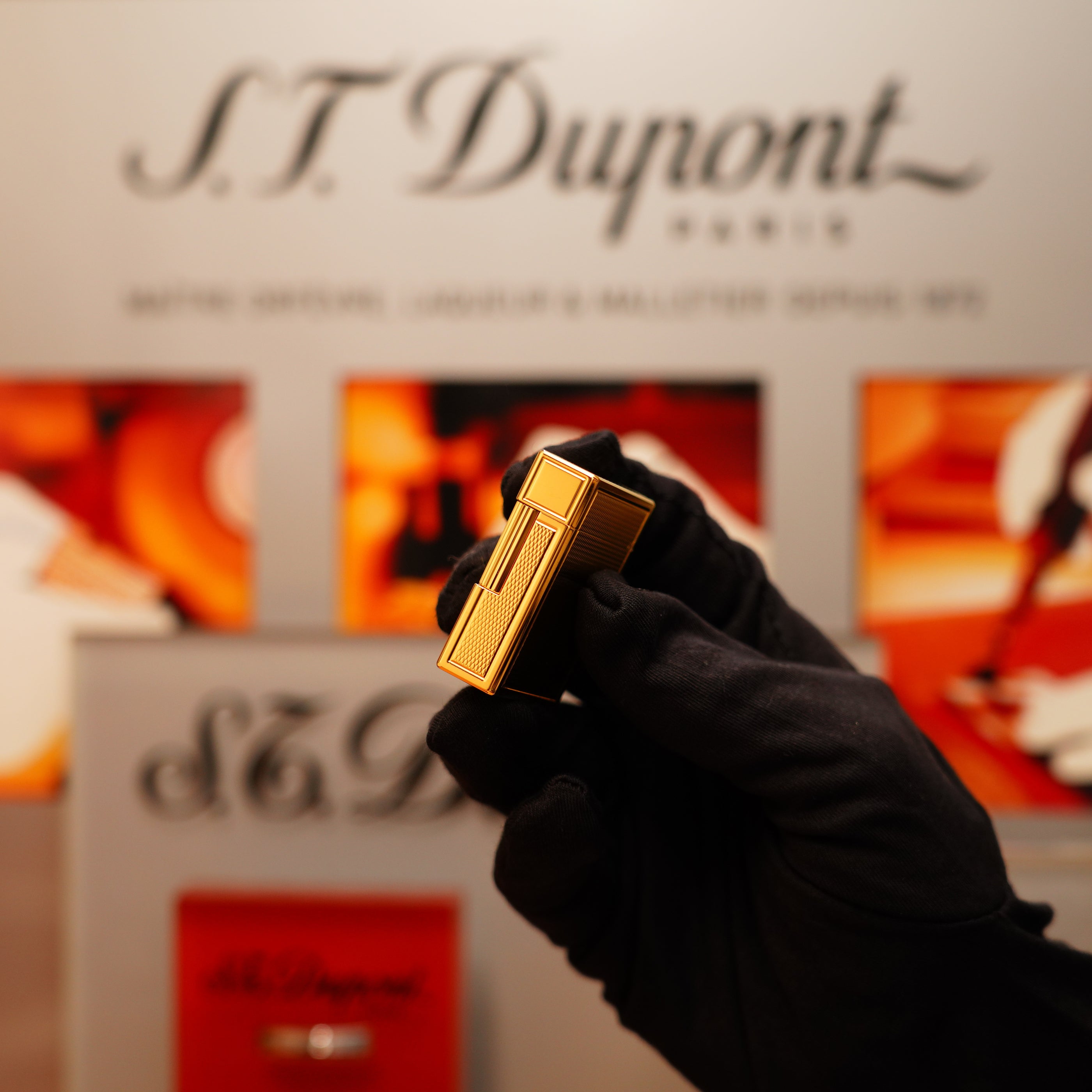 A gloved hand holds a Vintage 1996 S.T. Dupont 24k Small Grain d'Org Iconic Ligne 1 Heavy Finish Lighter against a blurred S.T. Dupont display background.