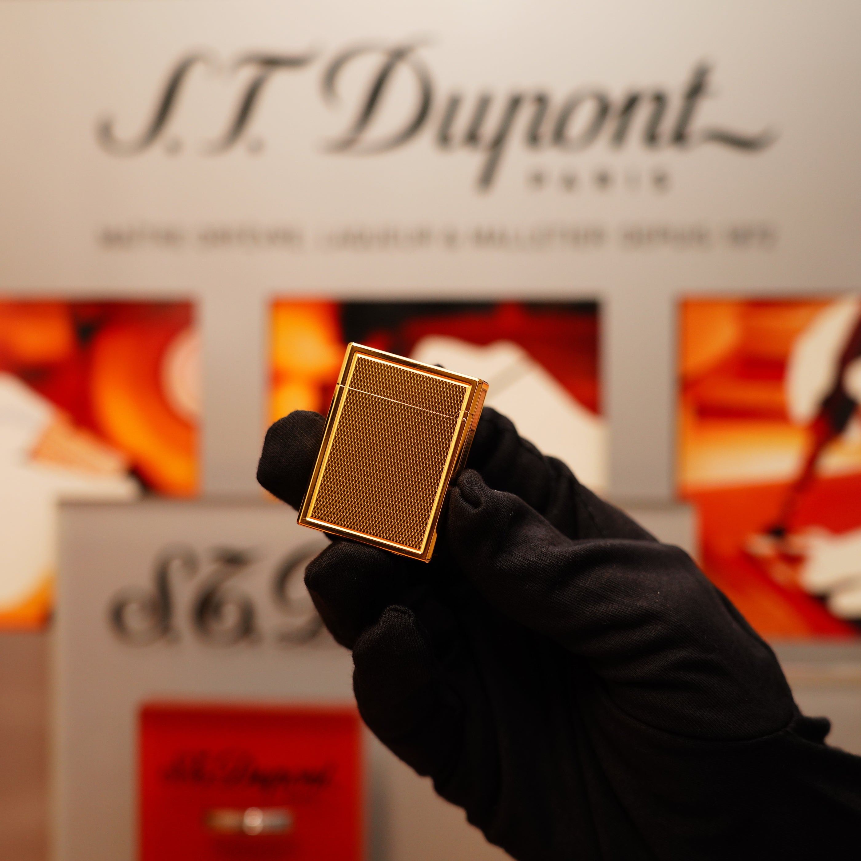 A gloved hand holds a Vintage 1996 S.T. Dupont 24k Small Grain d'Org Iconic Ligne 1 Heavy Finish Lighter in front of a blurred S.T. Dupont display.