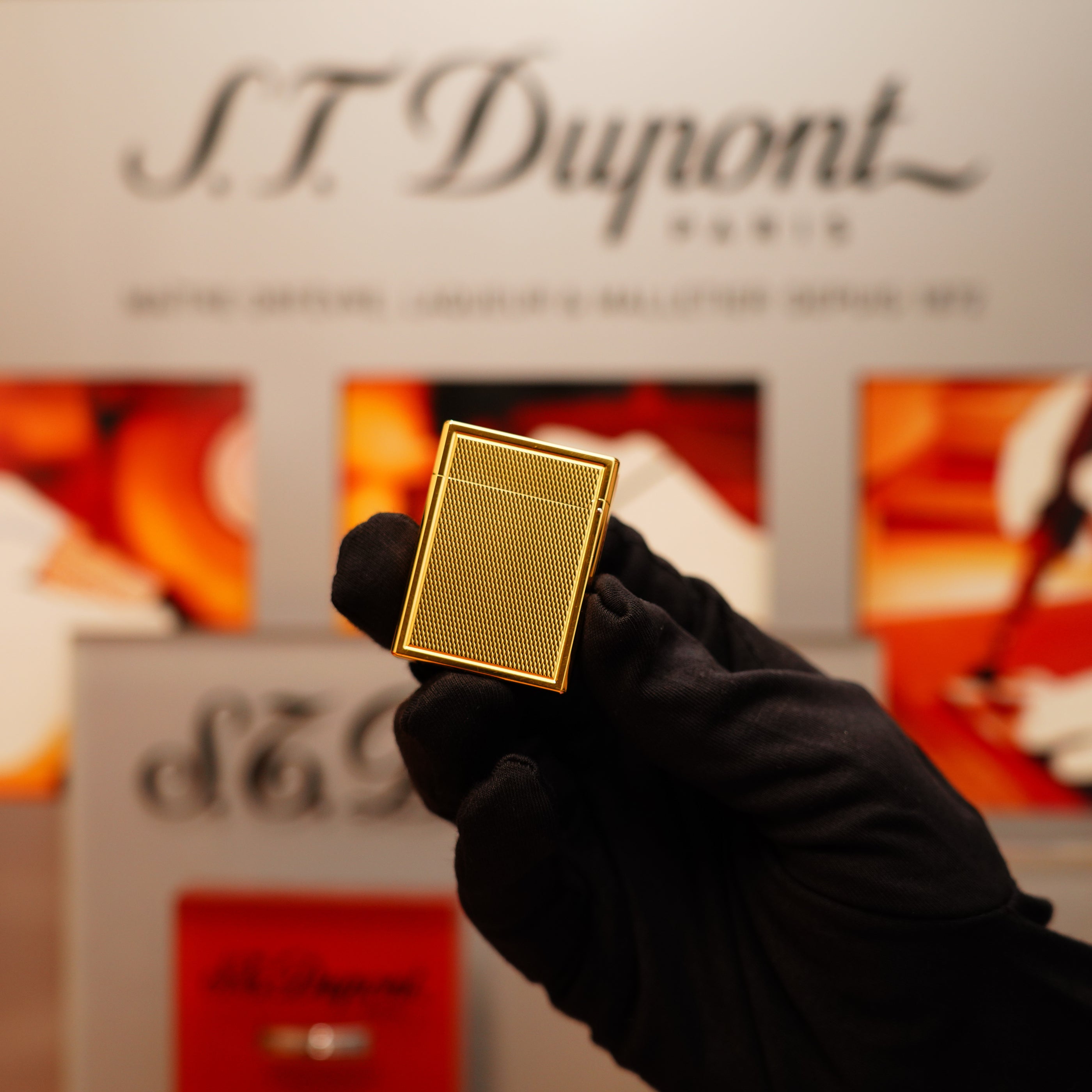 A gloved hand holds a gold rectangular lighter, identified as the Vintage 1996 St Dupont 24k Small Grain d'Org Iconic Ligne 1 Heavy Finish. The background showcases S.T. Dupont branding and an abstract design.