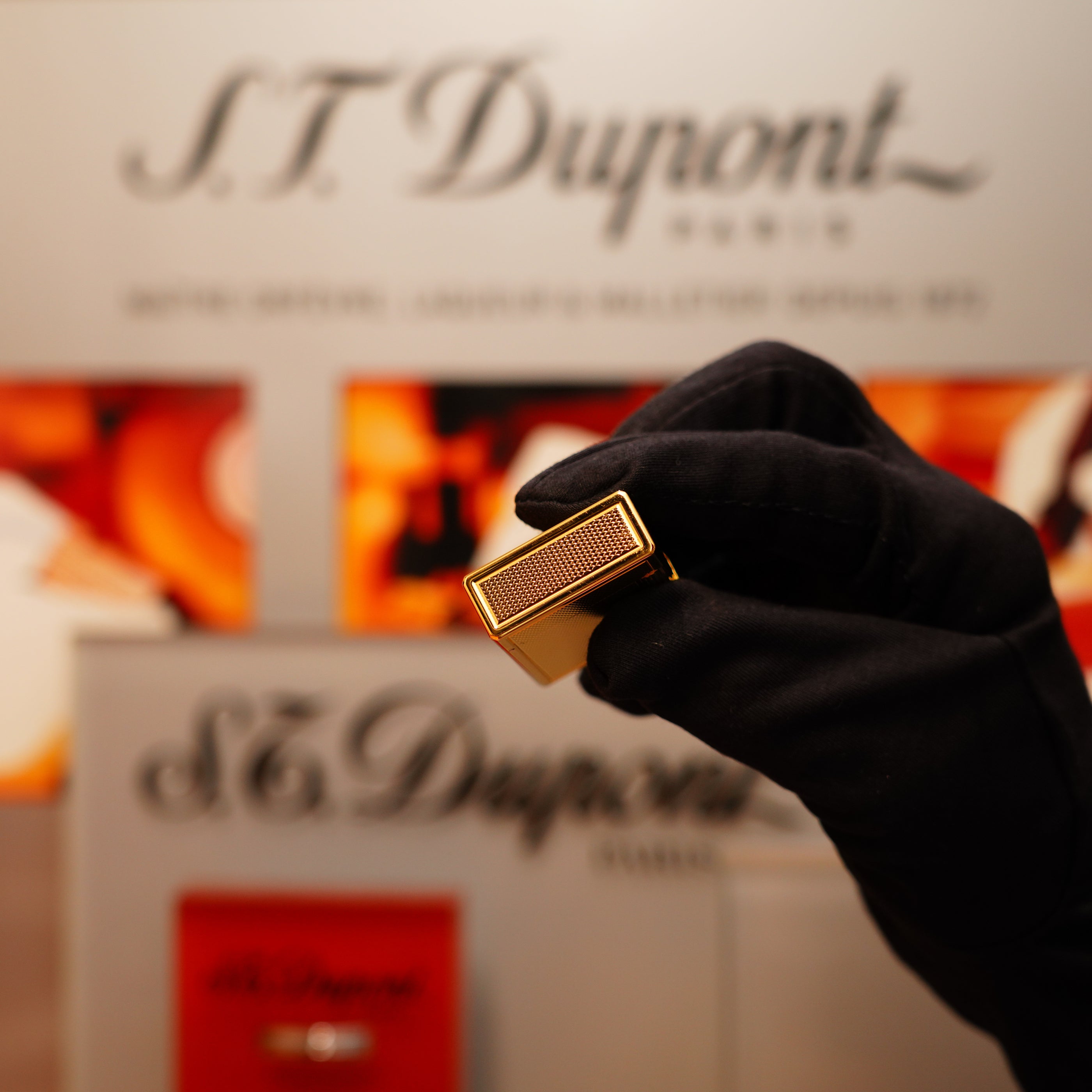 A gloved hand holds a Vintage 1996 S.T. Dupont 24k Small Grain d'Org Iconic Ligne 1 Heavy Finish Lighter against a blurred S.T. Dupont display.