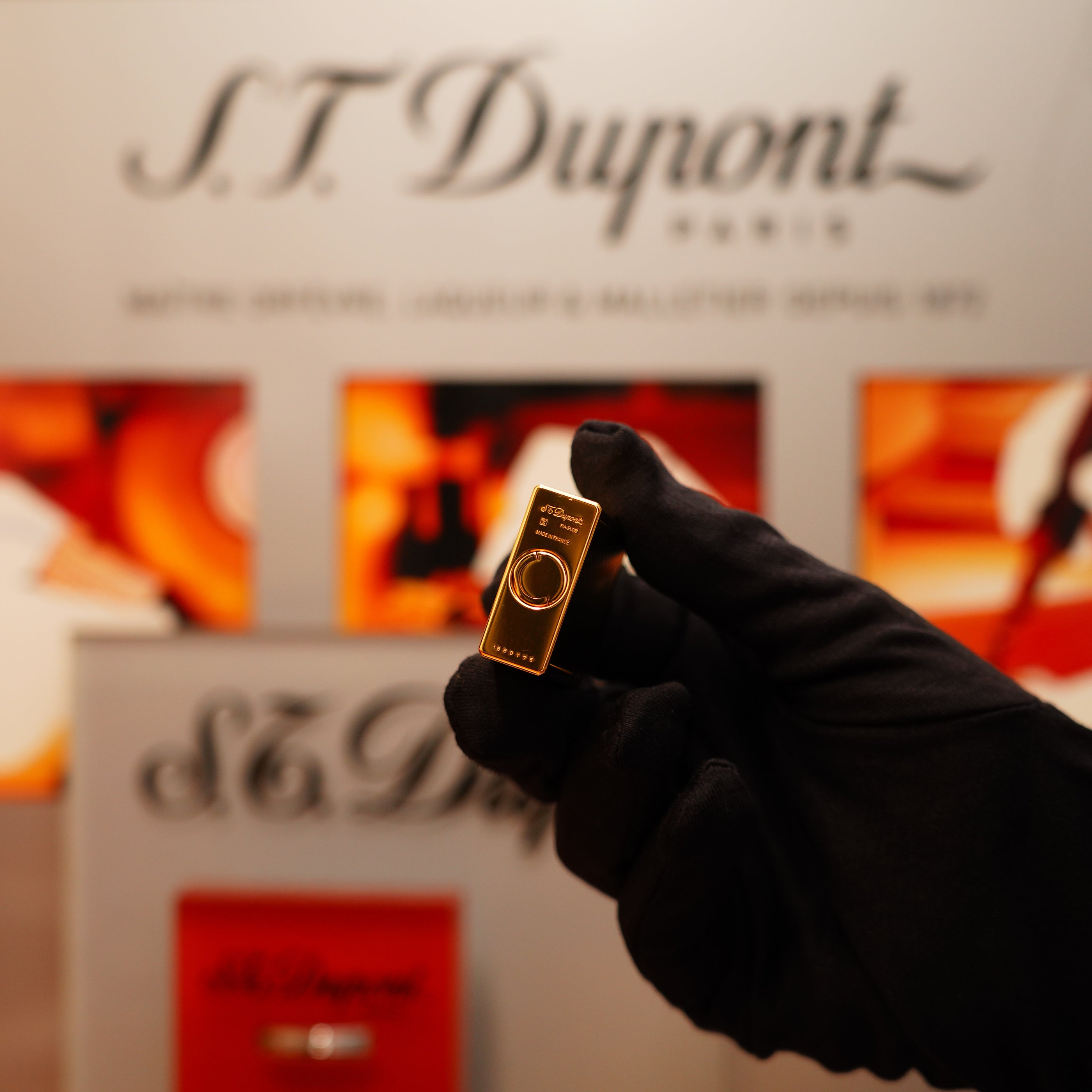 A gloved hand holds a Vintage 1996 St Dupont 24k Small Grain d'Org Iconic Ligne 1 Heavy Finish Lighter, with a blurred S.T. Dupont Paris sign in the background.
