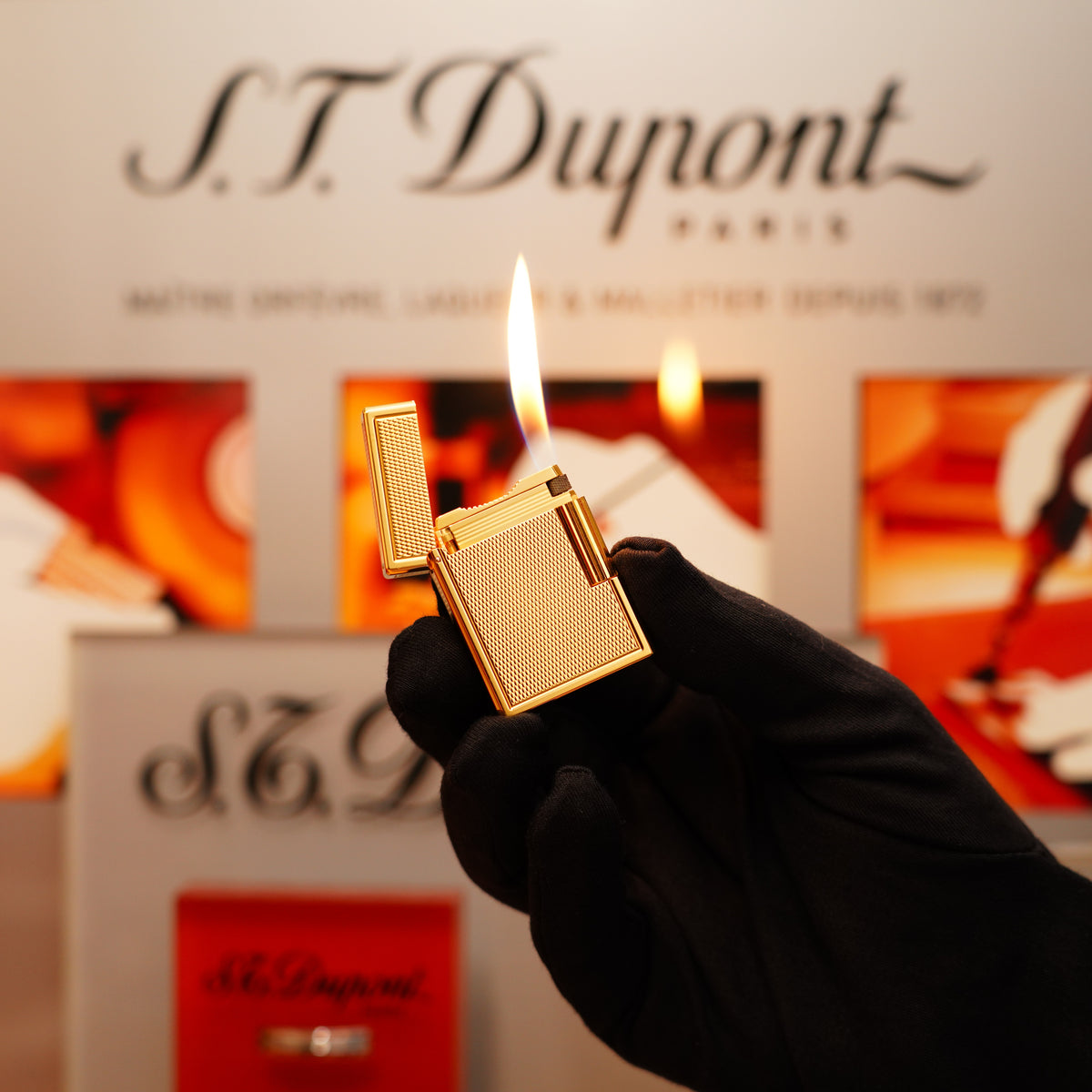 A gloved hand holds an open Vintage 1996 St Dupont 24k Small Grain d'Org Iconic Ligne 1 Heavy Finish Lighter with a visible flame. The background features S.T. Dupont branding and blurred images.