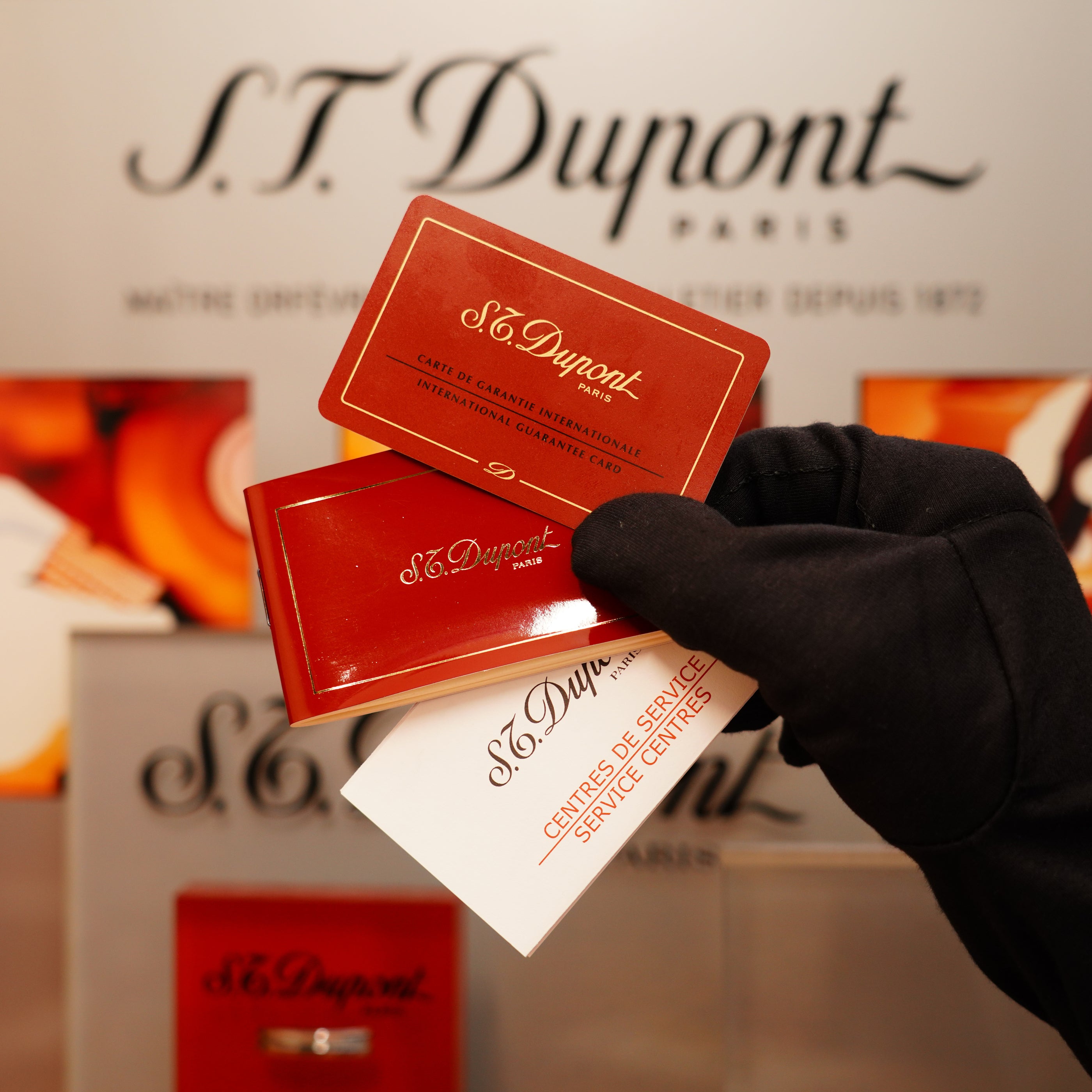 A gloved hand presents three S.T. Dupont cards against a blurred background showcasing the S.T. Dupont logo, related to the Vintage 1996 St Dupont 24k Small Grain d'Org Iconic Ligne 1 Heavy Finish Lighter.