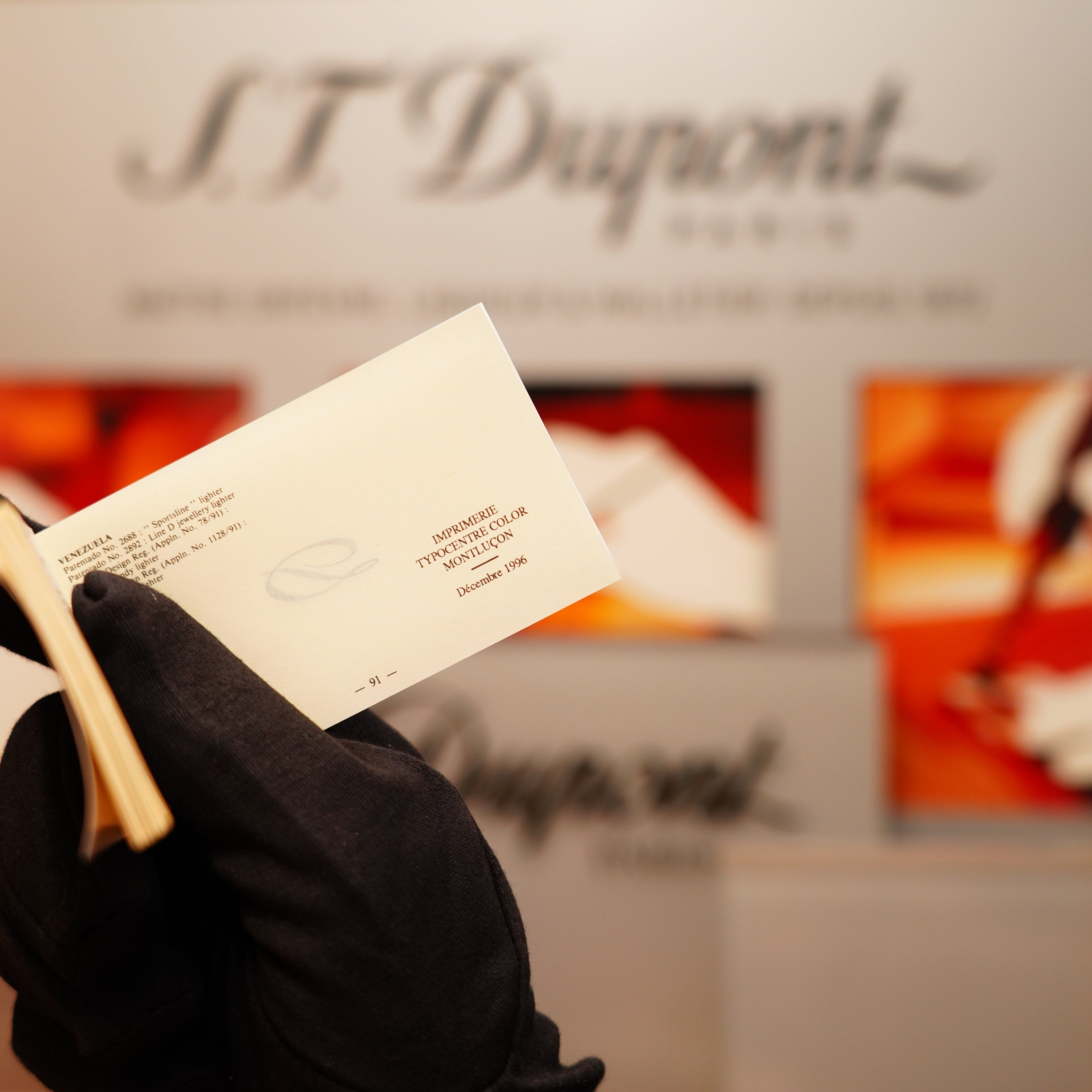 A gloved hand holds an invitation card with "S.T. Dupont" visible against a blurred luxury brand backdrop, reminiscent of the Vintage 1996 St Dupont 24k Small Grain d'Org Iconic Ligne 1 Heavy Finish Lighter.