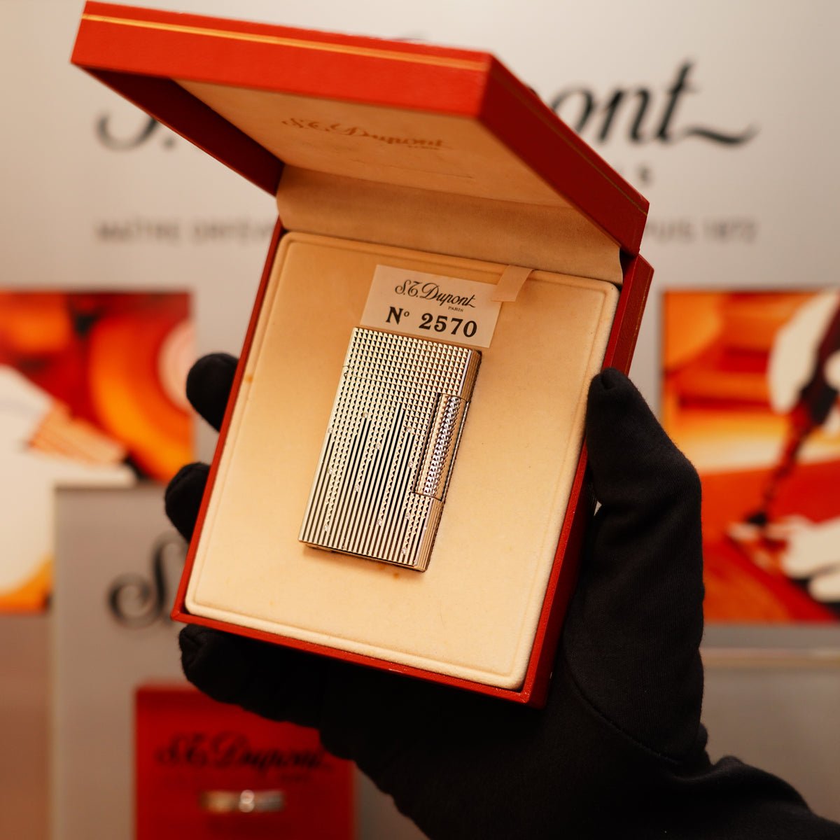 A gloved hand holds an S.T. Dupont Vintage 2005 Rare Natural Diamond Drops Large Ligne 2 Heavy Platinum Limited Edition Lighter in an open red box labeled "N° 2570" in an elegant store setting.