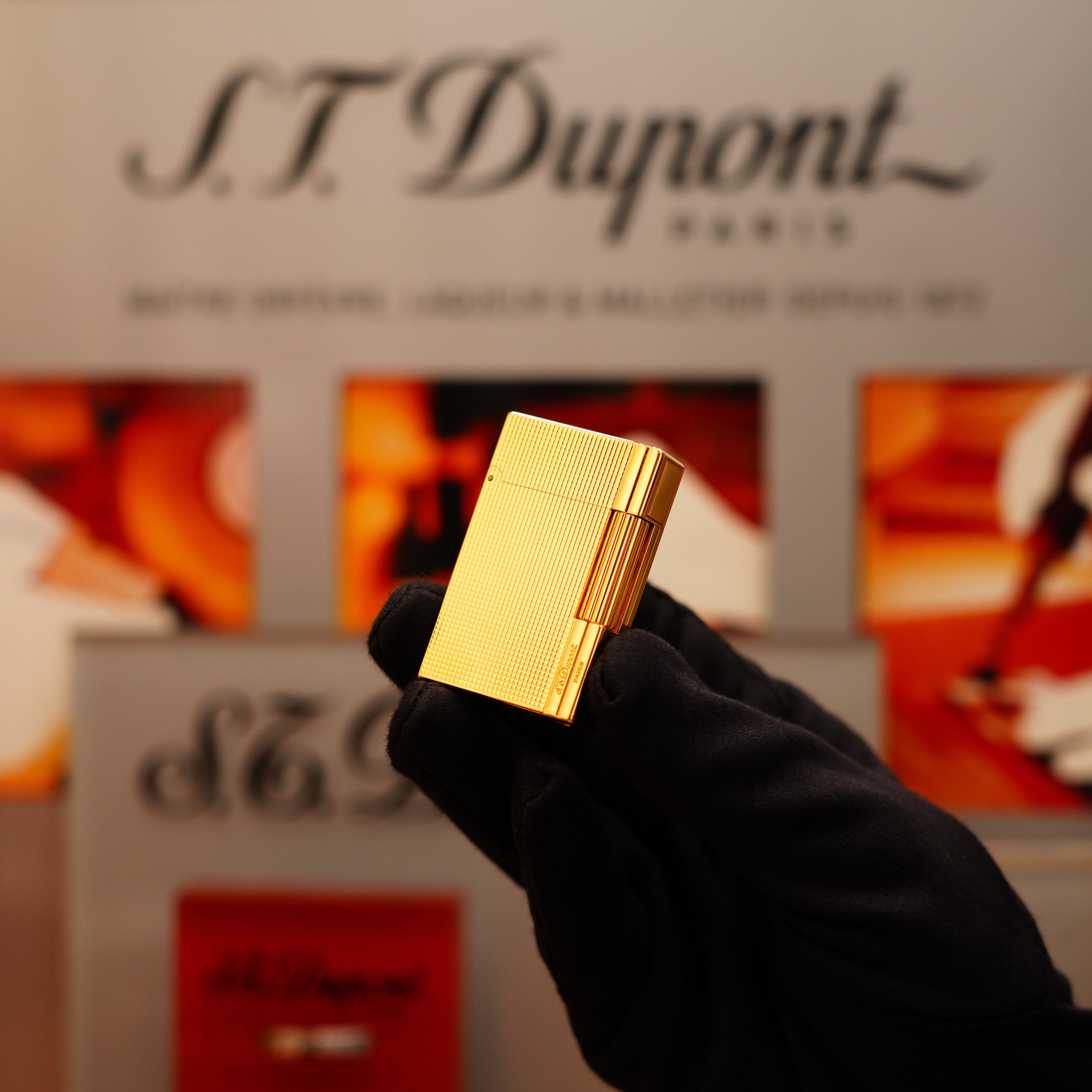 A person in a black glove holds a Vintage 2002 St Dupont Rare Heavy 24k Gold Finish Gatsby Vertical Line Pattern Ligne 2 lighter against a blurred background featuring S.T. Dupont branding.