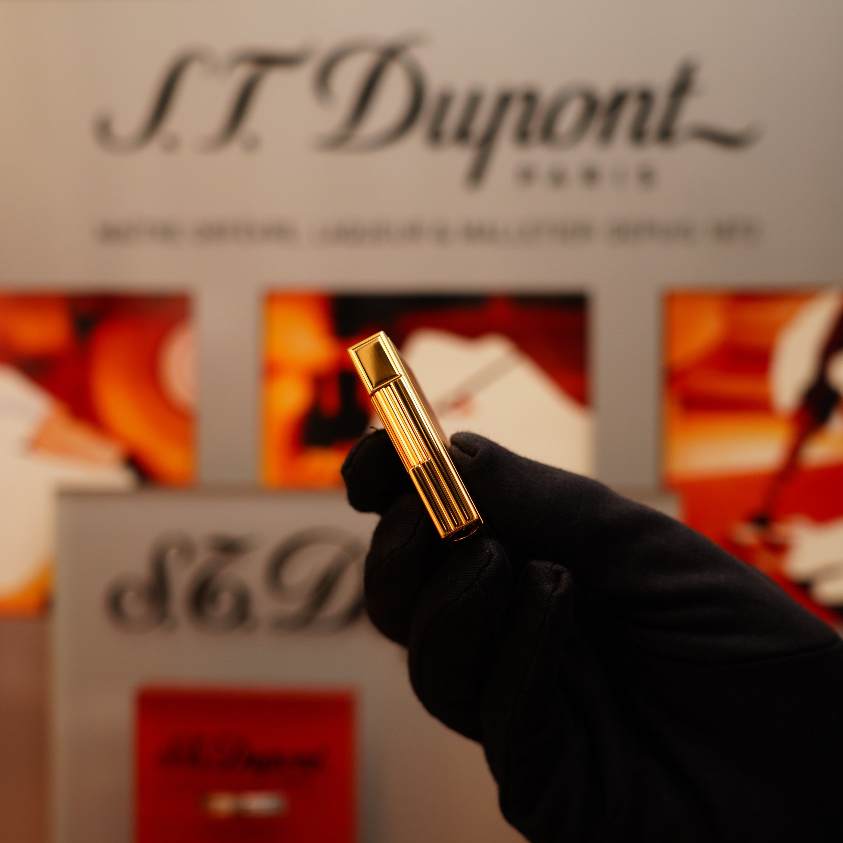 A gloved hand holds a Vintage 2002 S.T. Dupont Ligne 2 lighter with a 24k gold finish and Gatsby vertical line pattern against a blurred background featuring the brand's logo.