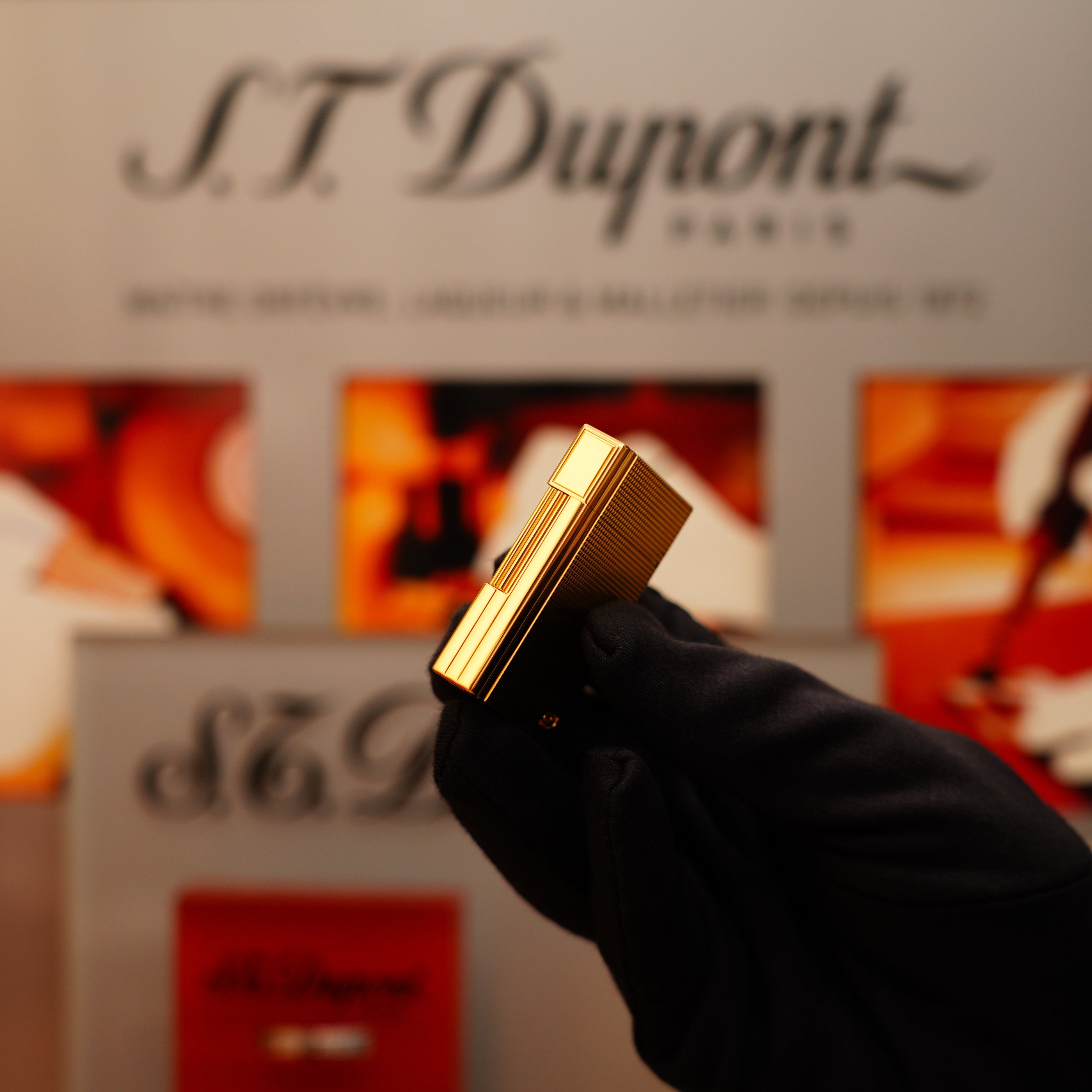 A gloved hand holds a Vintage 2002 St Dupont Rare Heavy 24k Gold Finish Gatsby Vertical Line Pattern Ligne 2 Lighter against a background featuring S.T. Dupont branding and blurred images.