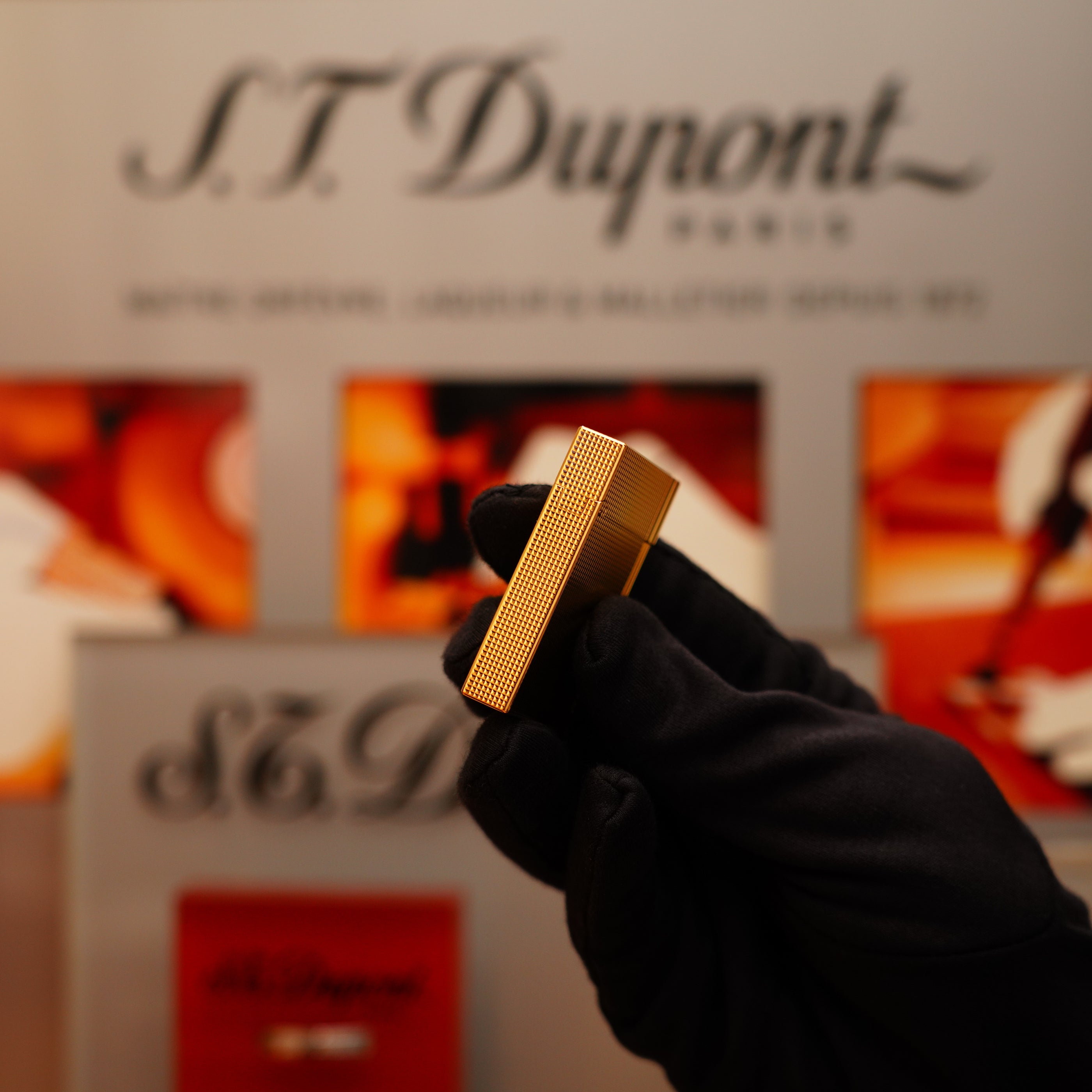 A gloved hand holds a Vintage 2002 S.T. Dupont Ligne 2 Lighter with a heavy 24k gold finish and Gatsby vertical line pattern, in front of a blurred branded background.