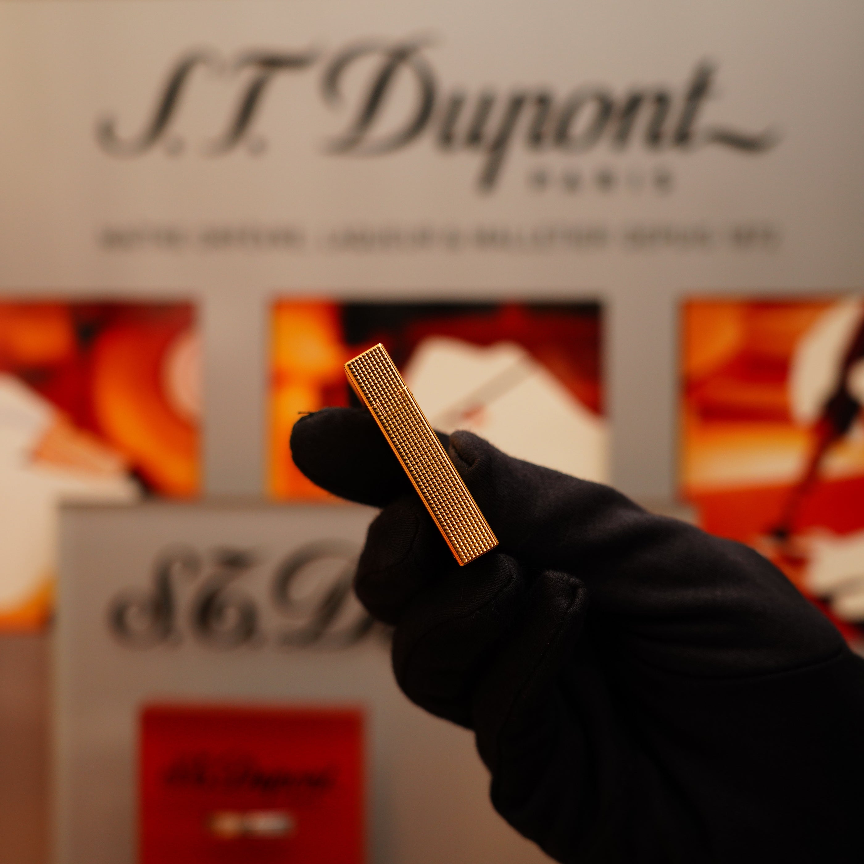 A gloved hand holds a Vintage 2002 S.T. Dupont Gatsby Ligne 2 lighter with a vertical line pattern and 24k gold finish in front of an S.T. Dupont Paris sign against a blurred, colorful background.