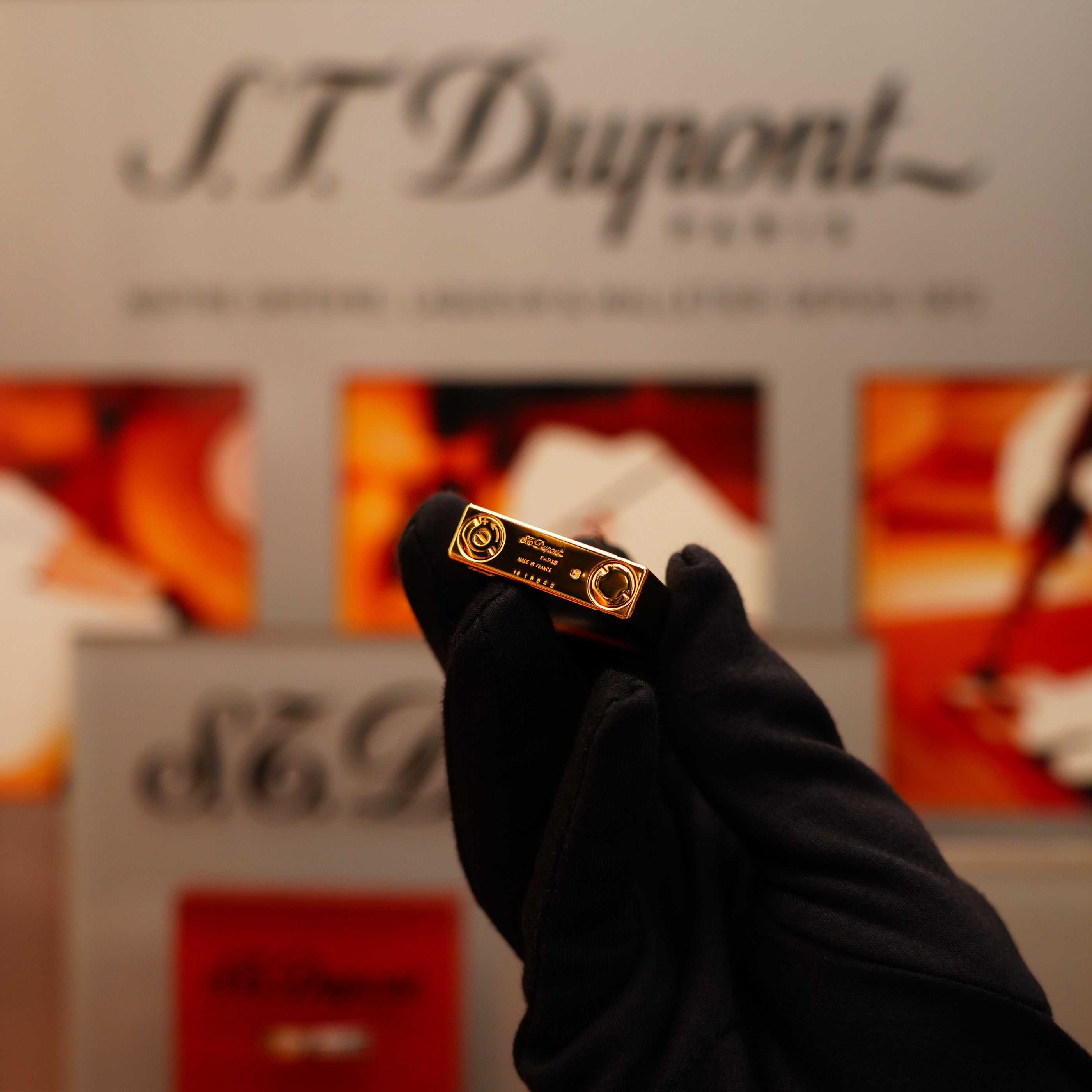 A gloved hand holds a Vintage 2002 St Dupont Rare Heavy 24k Gold Finish Gatsby Vertical Line Pattern Ligne 2 Lighter, with a blurred S.T. Dupont sign in the background.