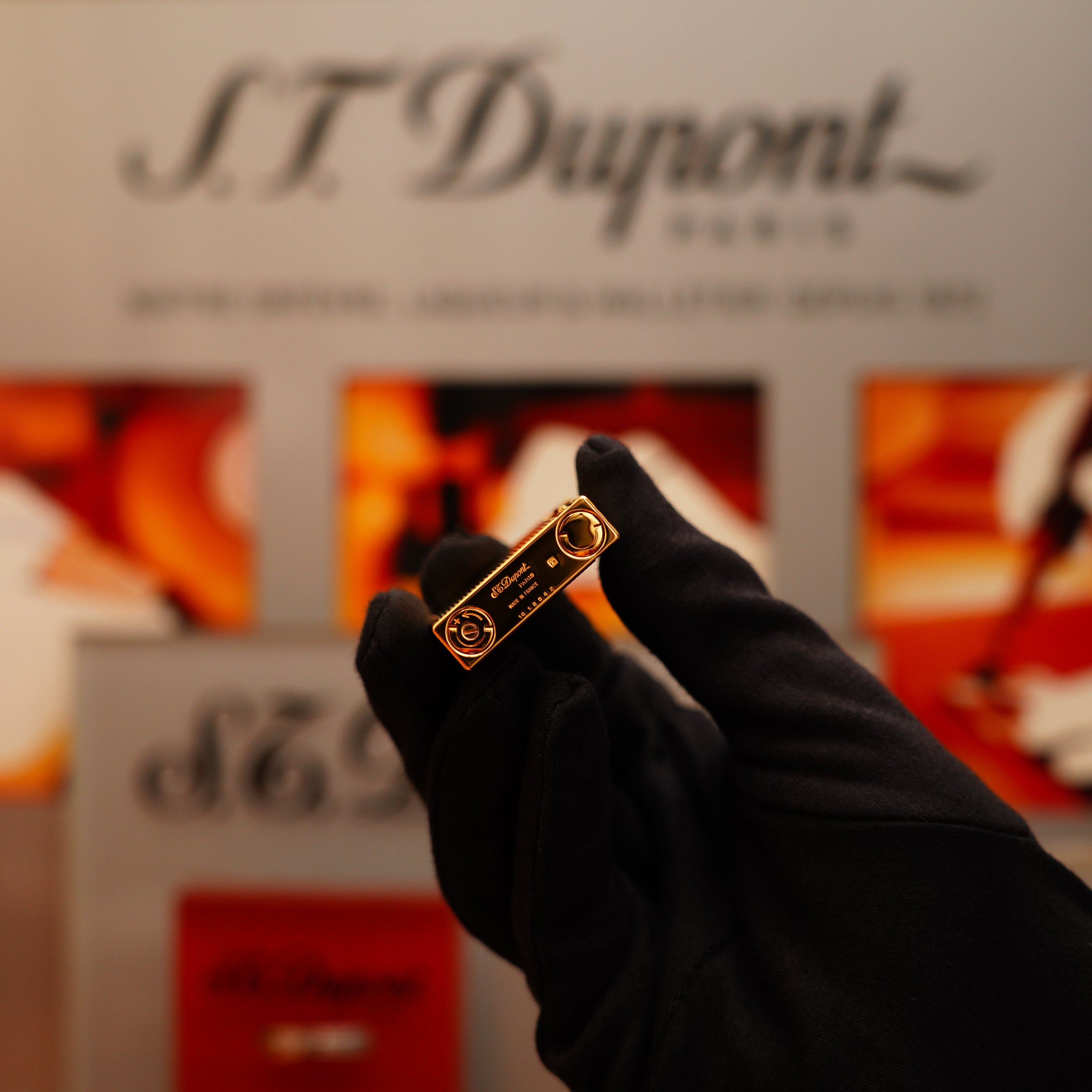 A gloved hand holds a Vintage 2002 S.T. Dupont Rare Heavy 24k Gold Finish Gatsby Ligne 2 Lighter with Vertical Line Pattern, while a blurred S.T. Dupont sign is visible in the background.