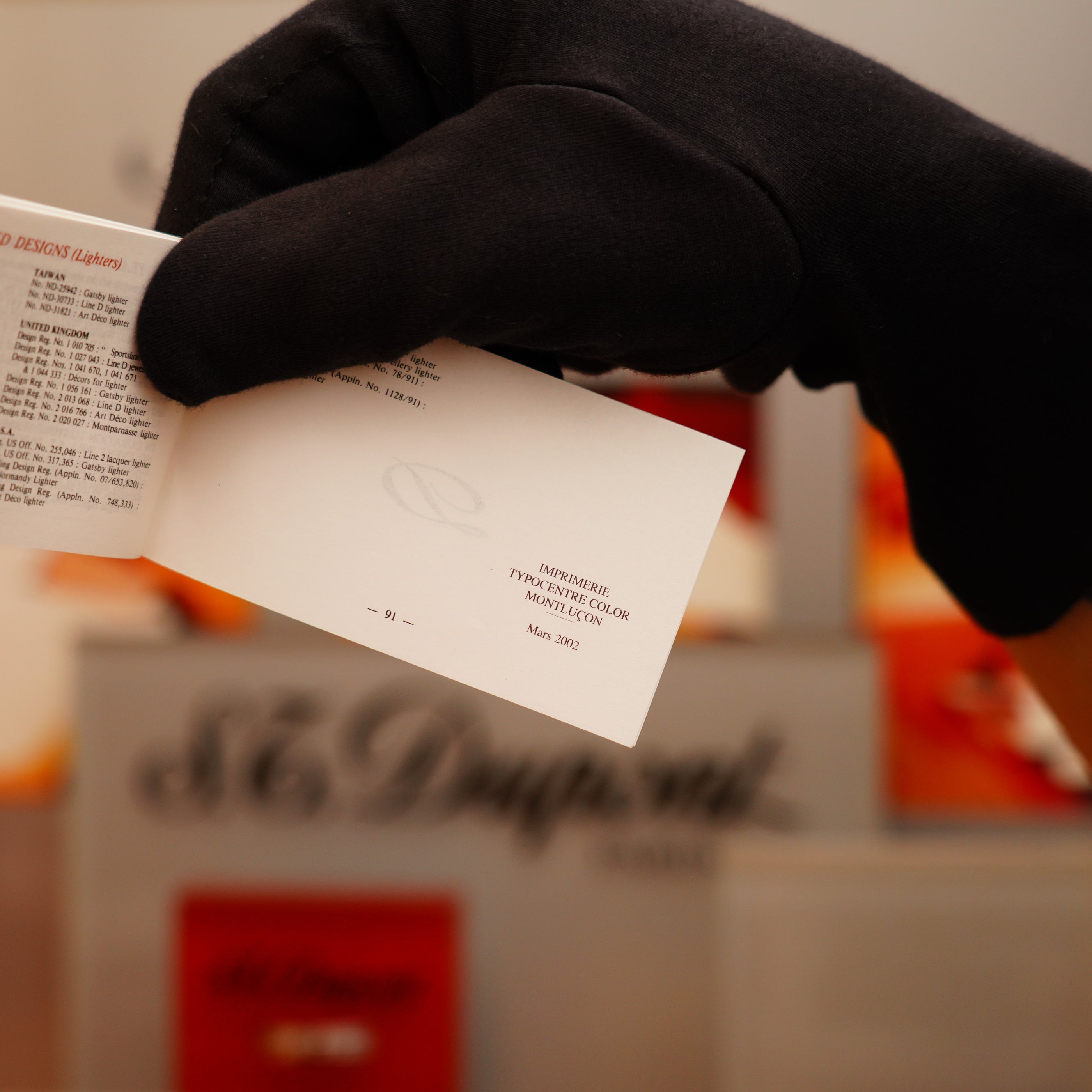 A gloved hand holds a small white card with text, against a blurred background featuring the S.T. Dupont brand name, known for products like the Vintage 2002 St Dupont Rare Heavy 24k Gold Finish Gatsby Vertical Line Pattern Ligne 2 Lighter.