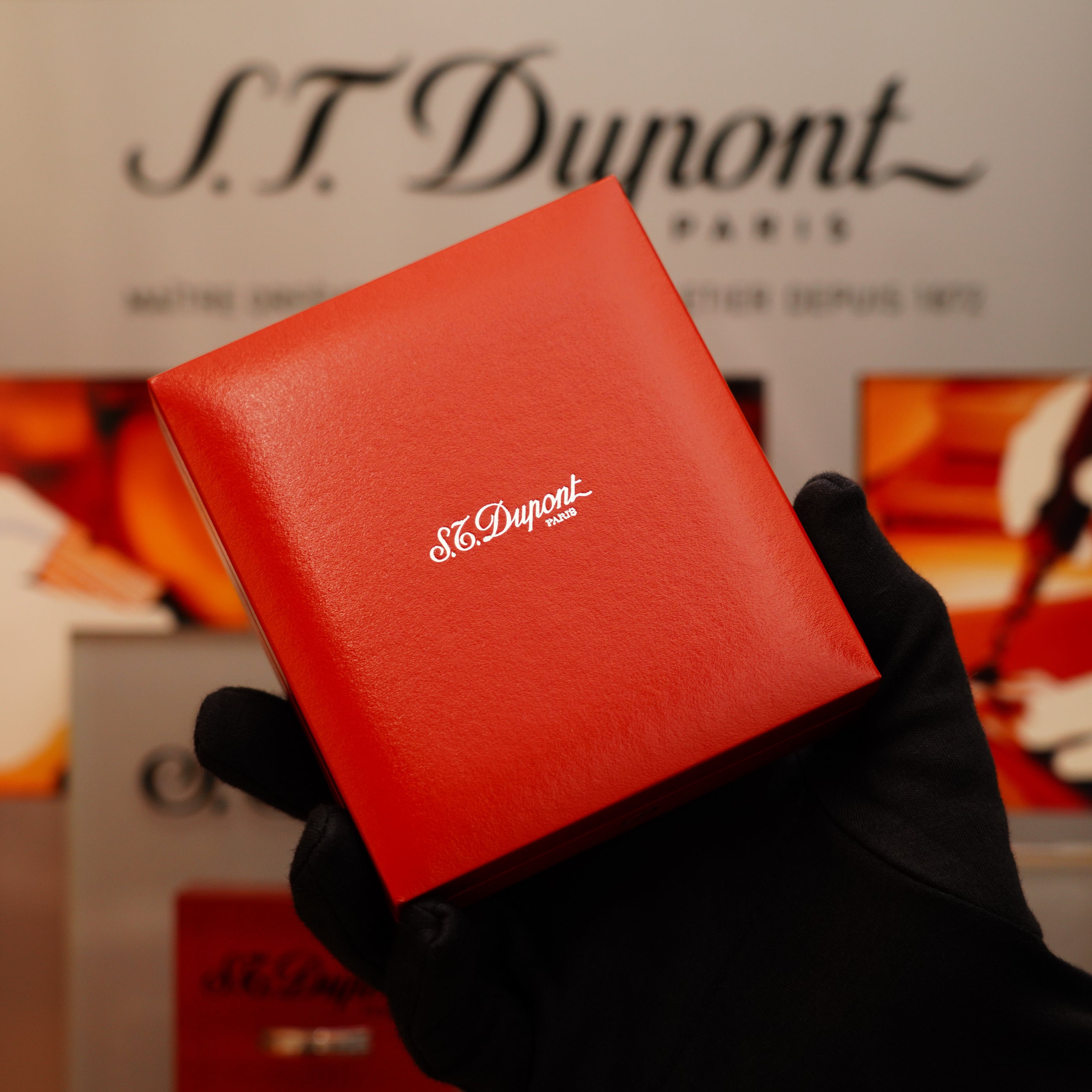 A gloved hand holds a red box containing the Vintage 2002 S.T. Dupont Ligne 2 Lighter with a 24k gold finish, set against a blurred S.T. Dupont sign.