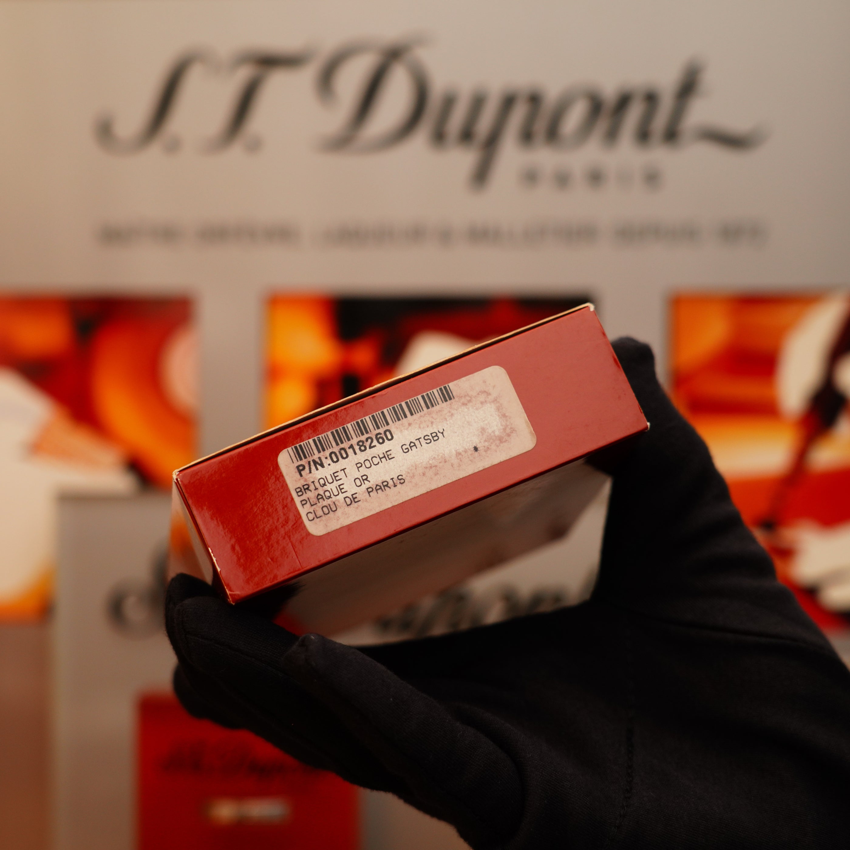 A gloved hand holds a red box with a barcode and label in front of a blurred "S.T. Dupont Paris" sign, showcasing the Vintage 2002 St Dupont Rare Heavy 24k Gold Finish Gatsby Vertical Line Pattern Ligne 2 Lighter.