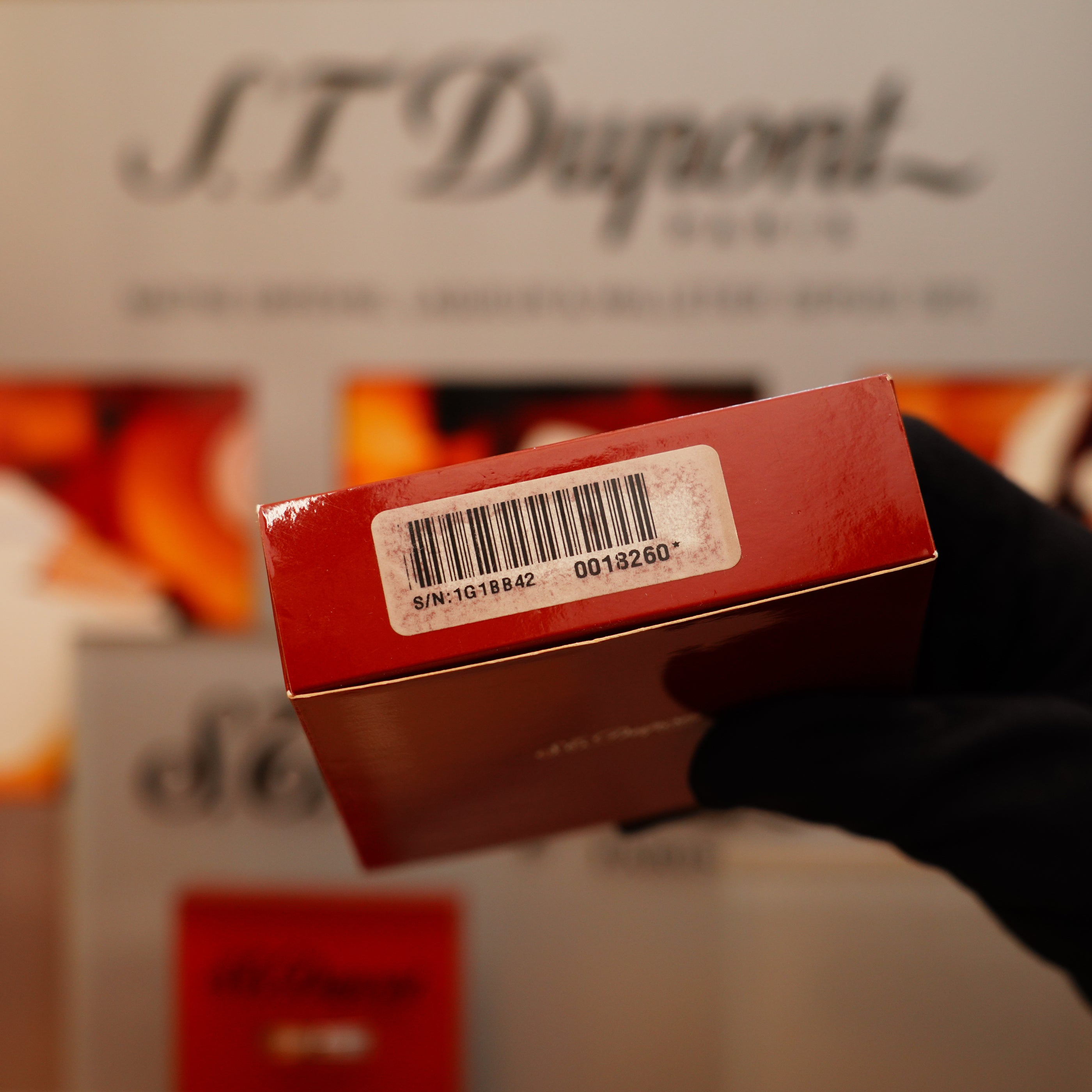 A gloved hand holds a red box with a barcode sticker and serial number. Blurred in the background is marketing material for the Vintage 2002 S.T. Dupont Rare Heavy 24k Gold Finish Gatsby Vertical Line Pattern Ligne 2 Lighter.